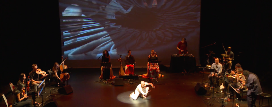 A dancer wearing all white in crouching in center of The Kitchen's performance space. A semi-circle of musicans surrounds the dancer. Behind them is a large-scale video projection.