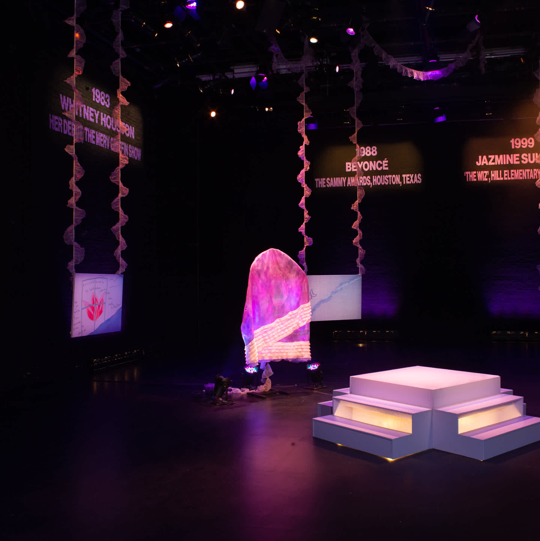 Illuminated periwinkle stage surrounded by three hanging digital drawings with light pink ruffles, a glowing bag sculpture and hand-painted text on the wall that reads left to right “1978 Diana Ross, ‘The Wiz,’ Motown Film, "1983 Whitney Houston, Her Debut, The Merv Griffin Show," "1988 Beyonce, The Sammy Awards, Texas," and "1999 Jazmine Sullivan, 'The Wiz,' Hill Elementary, Philadelphia."