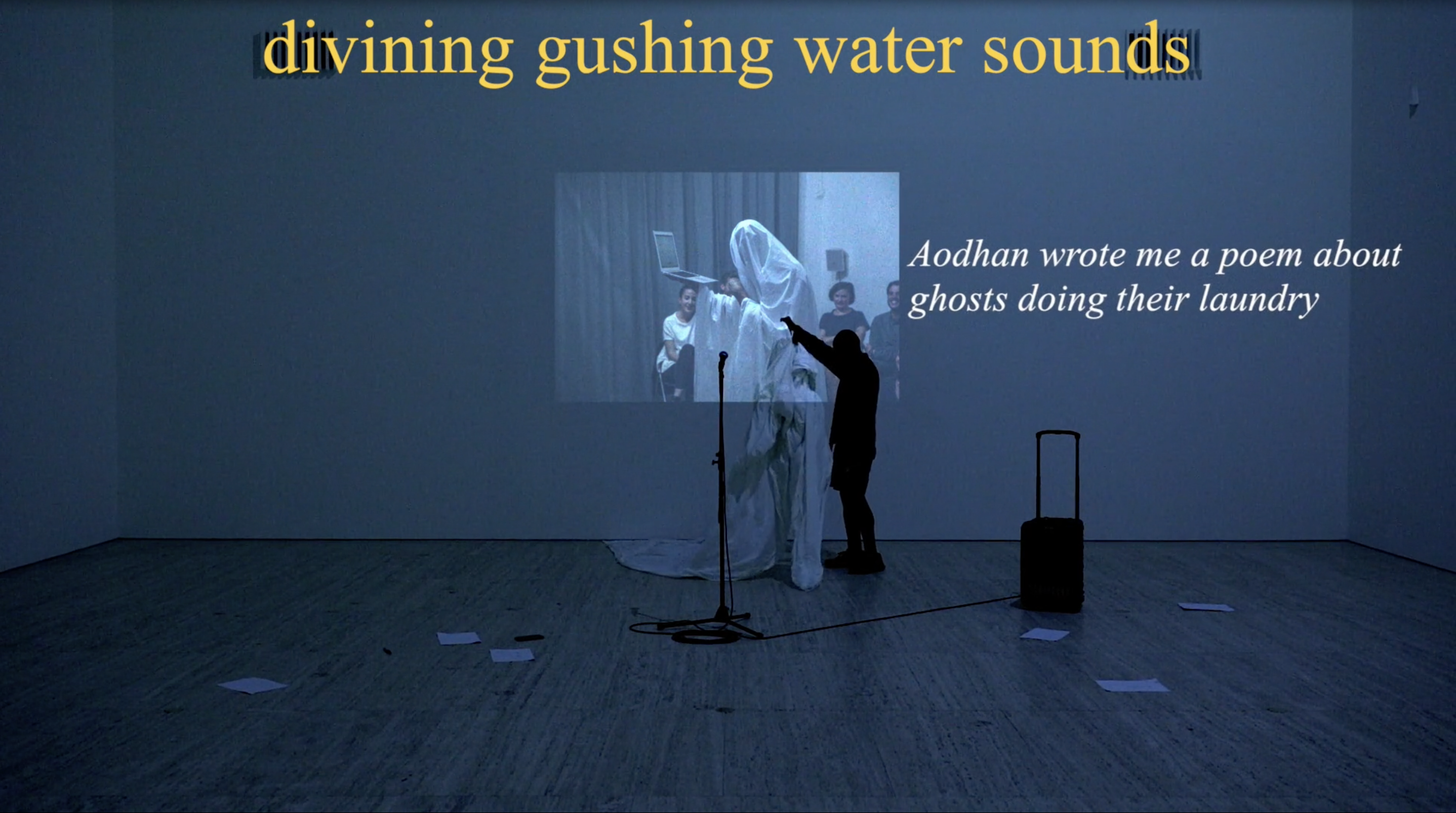 In a dark room with a video projection on the rear wall, Brian Fuata stands near a mic stands and handles a white sheet. Yellow text reading "diving gushing water sounds" appears at the top of the image. To the right of Brian, white, italicized text reads, "Aodhan wrote me a poem about ghosts doing their laundry."