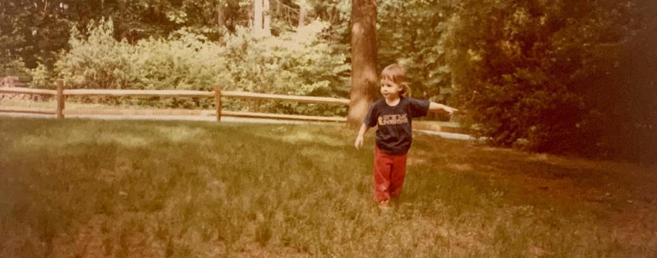 A small child stands on a lawn with a split beam fence and trees behind them. The child looks left while pointing to their right.