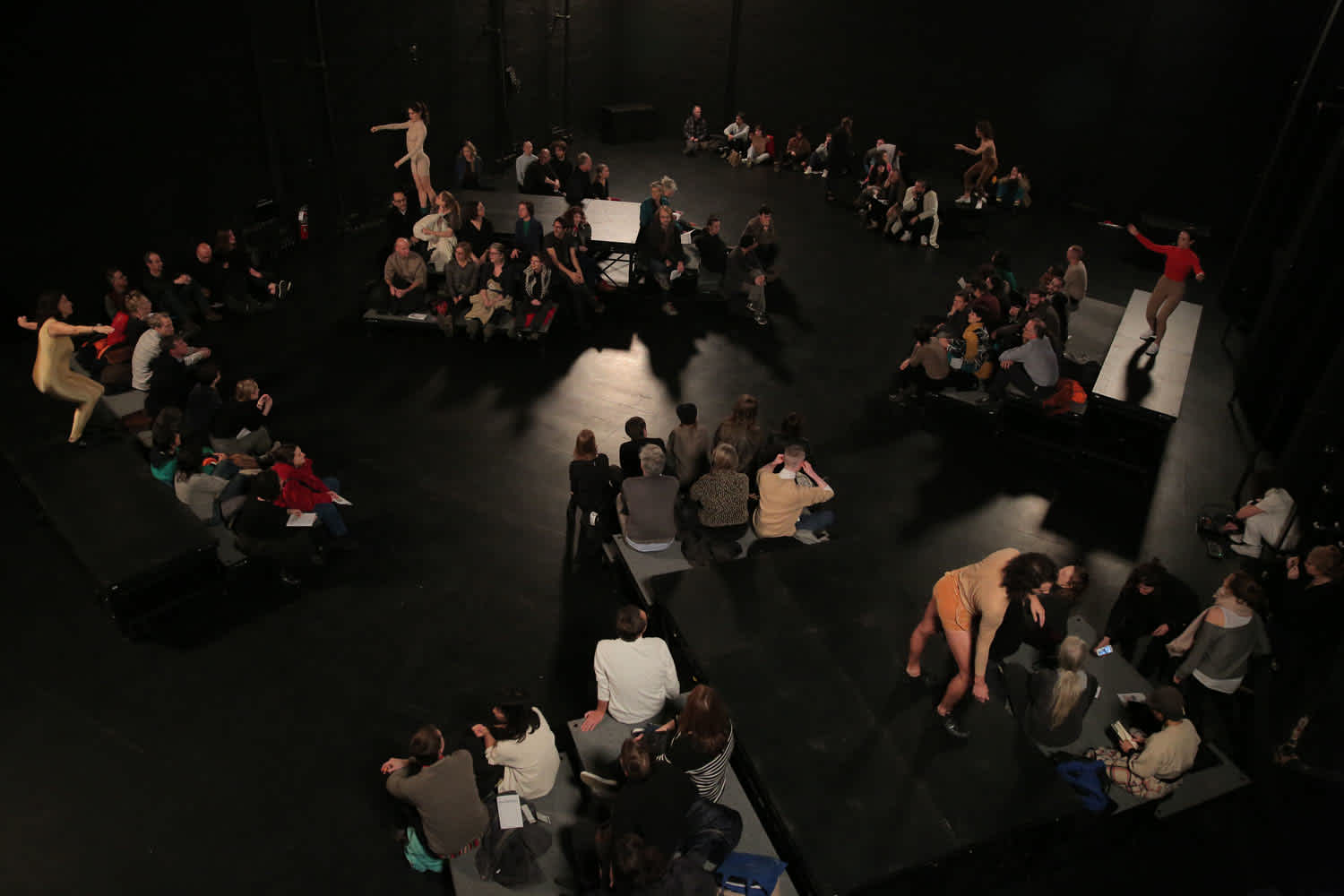 An aerial view of the performance space for Moriah Evans' work Configure. The audience is seated on platforms dispersed around the room. Performers are dispersed among the audience and some are standing on top of the platforms.