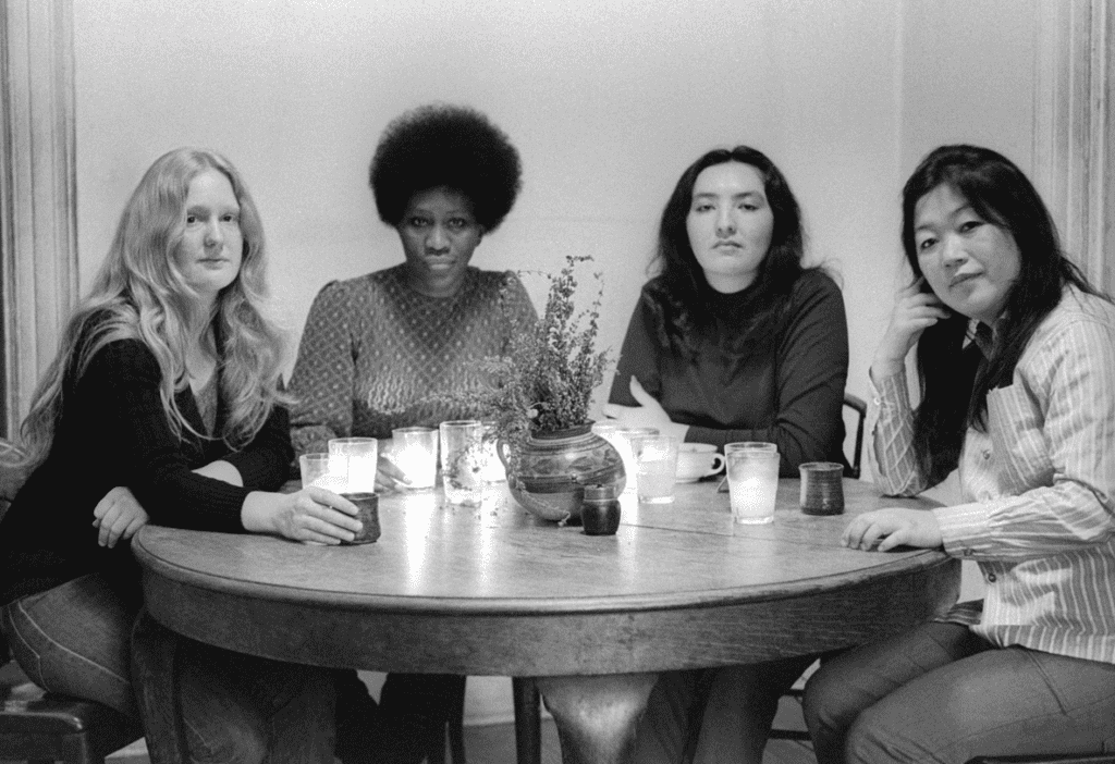A horizontal black and white photograph depicting Mary Lucier, Charlotte Warren, Cecilia Sandoval, and Shigeko Kubota facing directly and turning toward the camera sitting next to each other on a round dining table with various lit candles and a floral arrangement in a vase interspersed around them. 