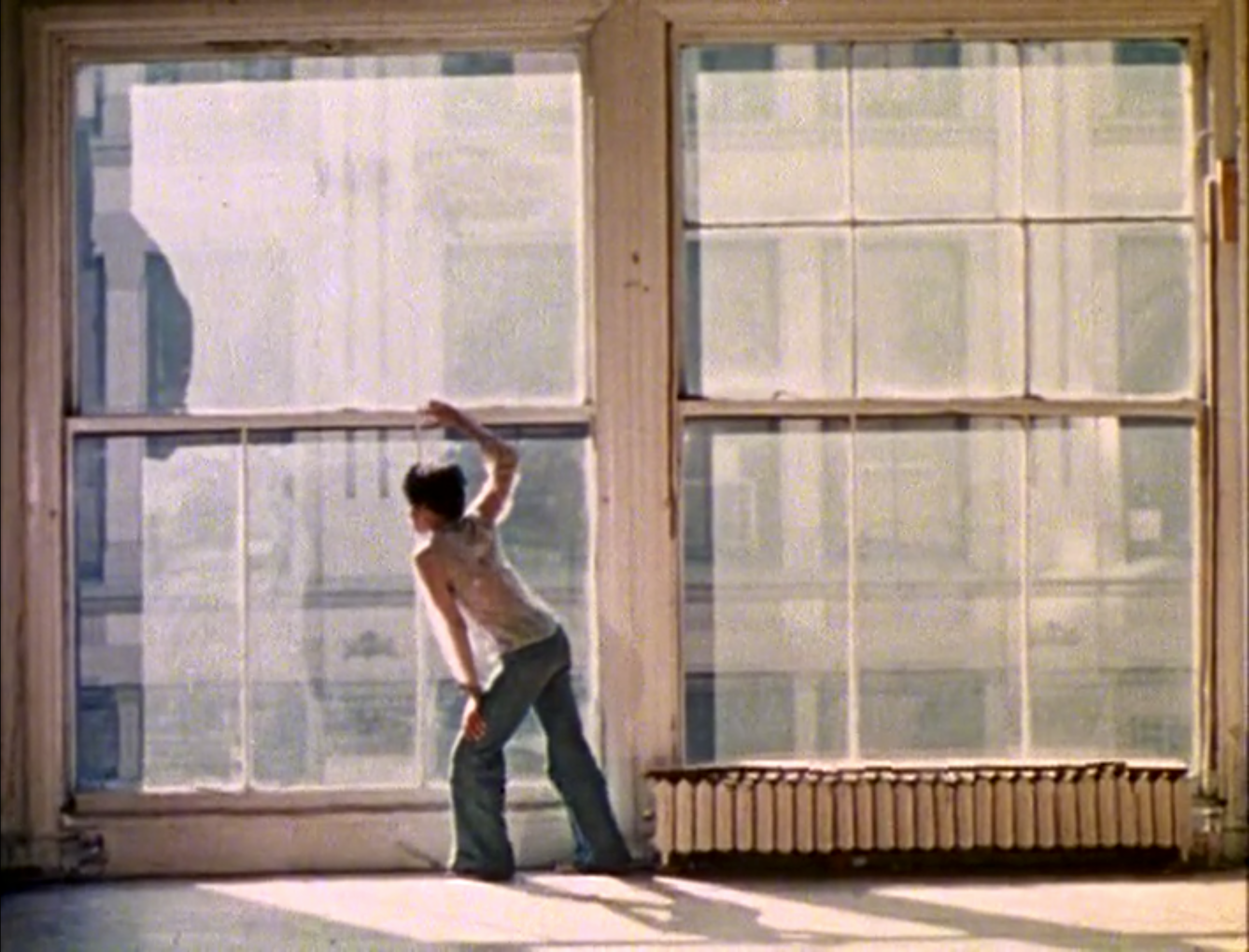A dancer stands in a sun-lit loft space in front of two large windows and looks out towards the left.