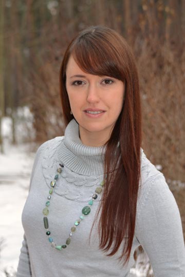 Karina Sumner-Smith standing in front of some tree's wearing a grey sweater and a green necklace.