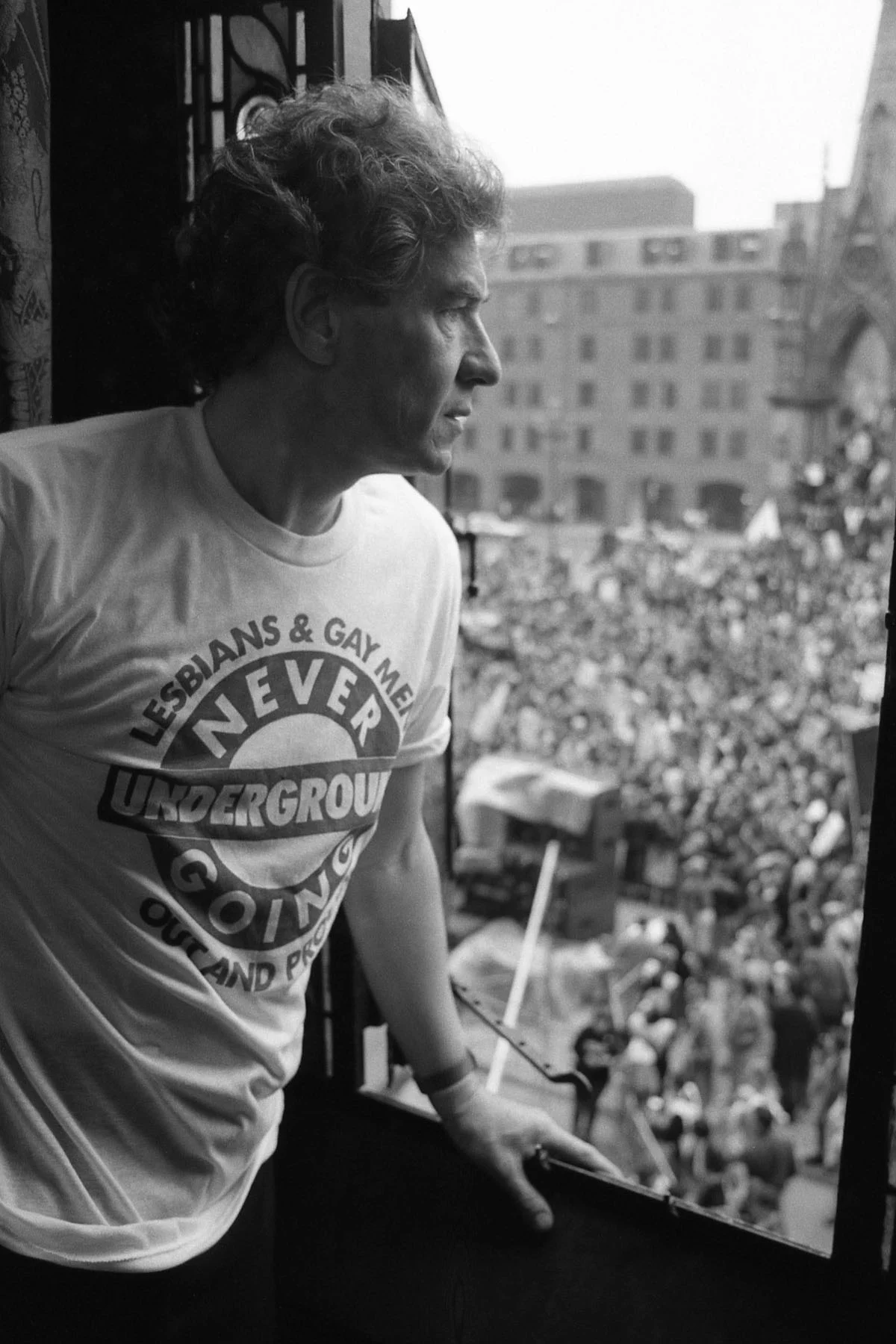A black-and-white photograph of English actor Sir Ian McKellen standing by an open window, looking out at a demonstration taking place in Manchester. Hundreds of protestors are visible below him.