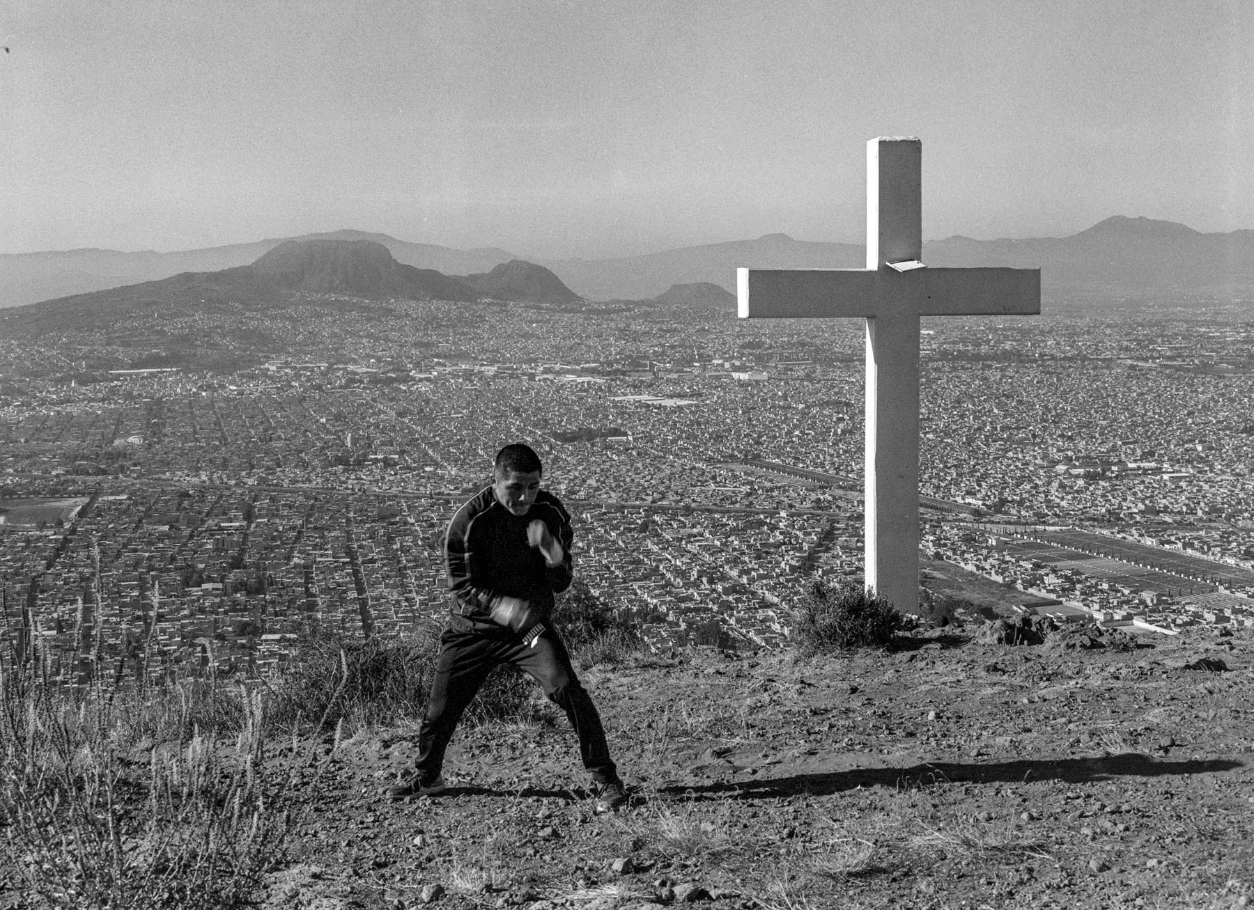 A black-and-white photograph of a boxer shadow boxing in front of a cross that overlooks his hometown of Chimalhuacán, Mexico.
