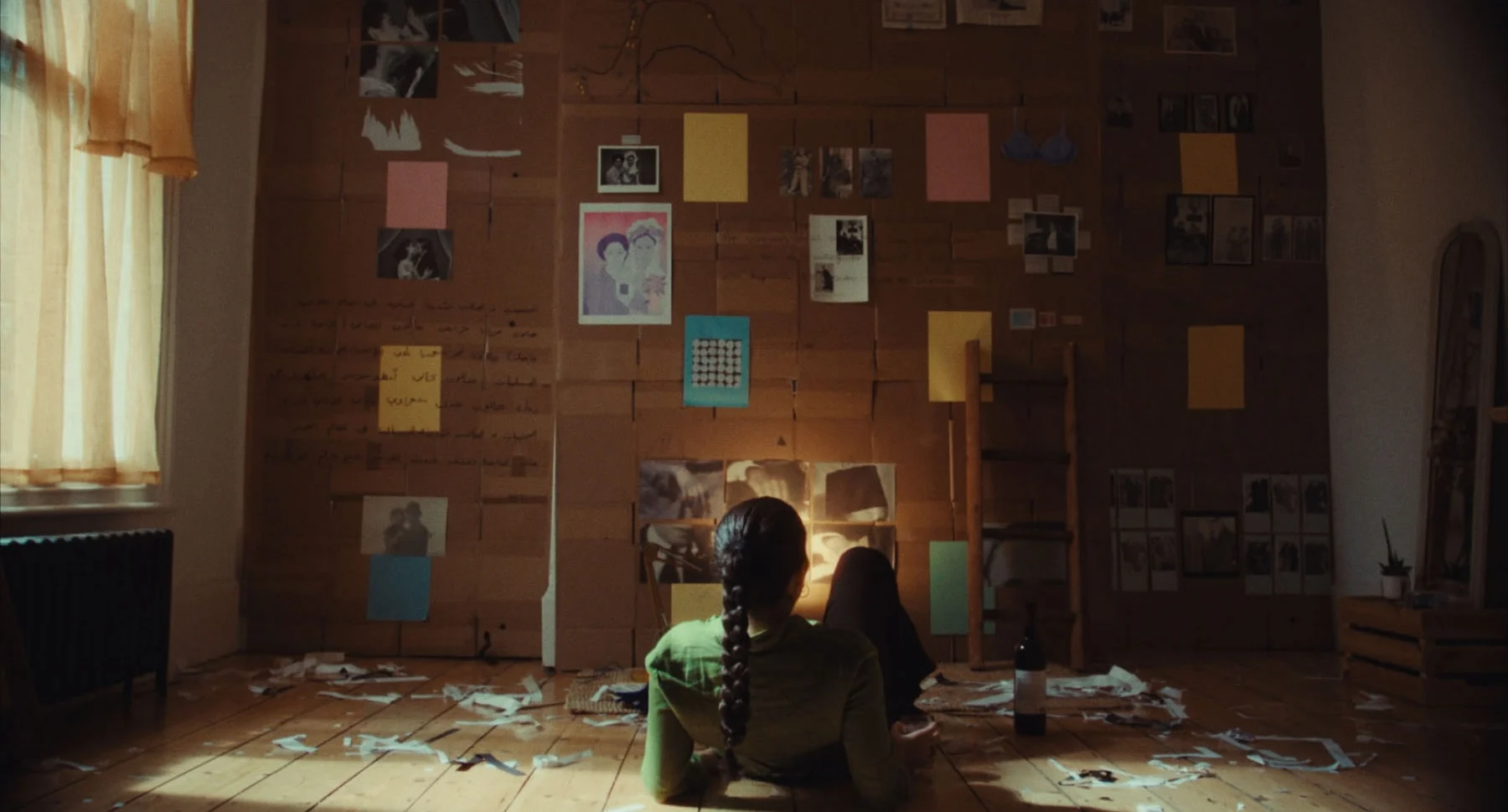 A still from the film Neo Nahda by May Ziade. The still shows a young woman sitting on the floor of her very messy room. One of her walls is fully covered by cardboard and miscellaneous “evidence” of Arab queer and feminist photographic histories of the early 20th century. 
