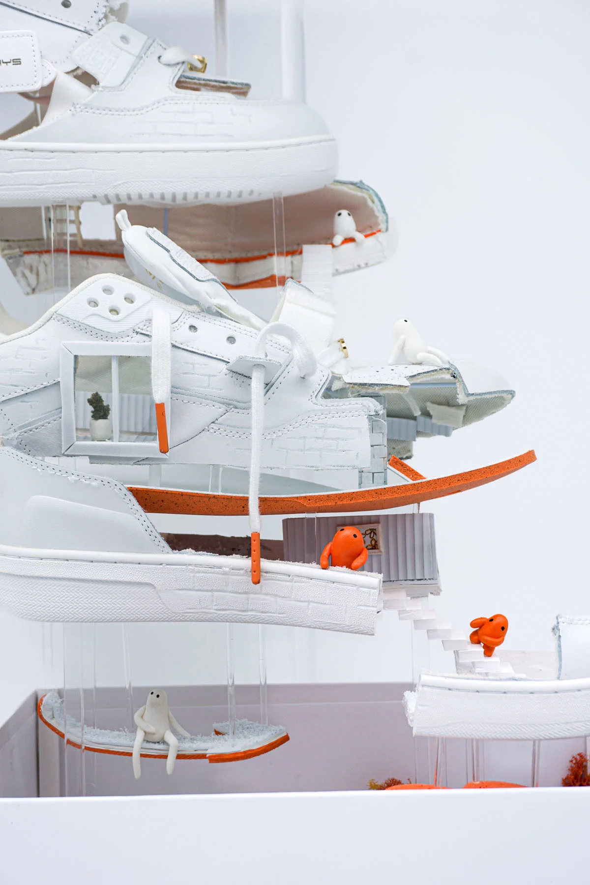 A close up photograph of a sculpture involving a sneaker separated into vertical layers to create an apartment. Inside the apartment are small orange figures, a pool, garden and stairs between each floor. In the photo you see a close up of the window in one of the floors, a small art piece in one of the rooms and the figures living in the space. 
