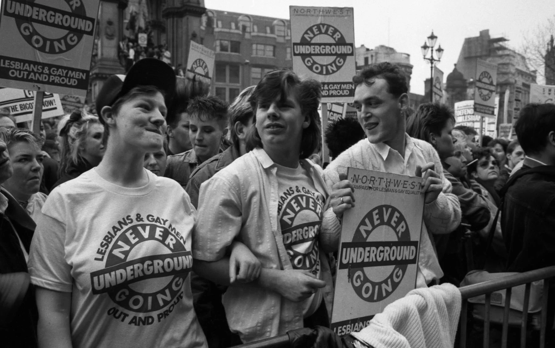 A black-and-white photograph of three smiling protestors standing next to each other behind a metal barricade, holding signs that read “LESBIANS & GAY MEN NEVER GOING UNDERGROUND OUT AND PROUD”. Behind them stand dozens of other protestors, holding signs with the “NEVER GOING UNDERGROUND” logo on them.