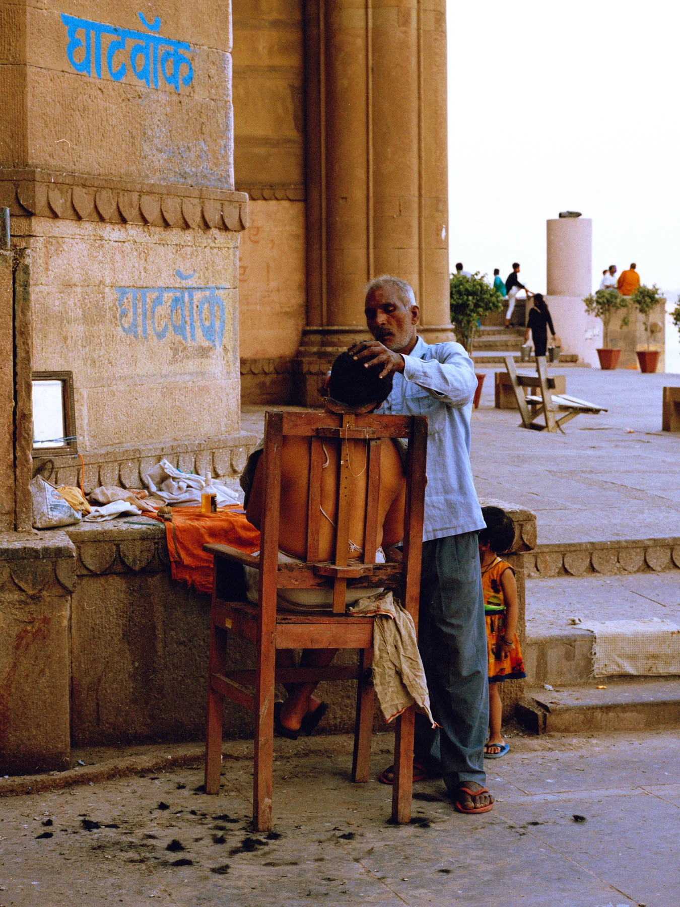 A photograph of a middle-aged barber on the ‘ghats’ of Varanasi, shaving the beard of another man who is sitting on a wooden chair.