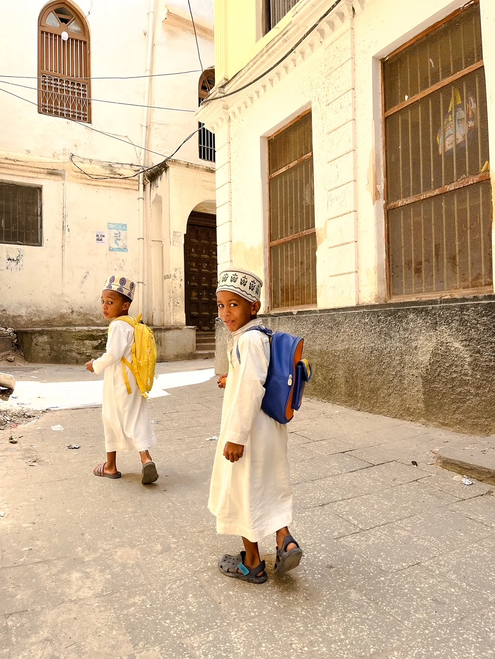 A photograph of two boys walking along a street in Zanzibar, Tanzania. They both glance back at the camera as they walk, smiling. 