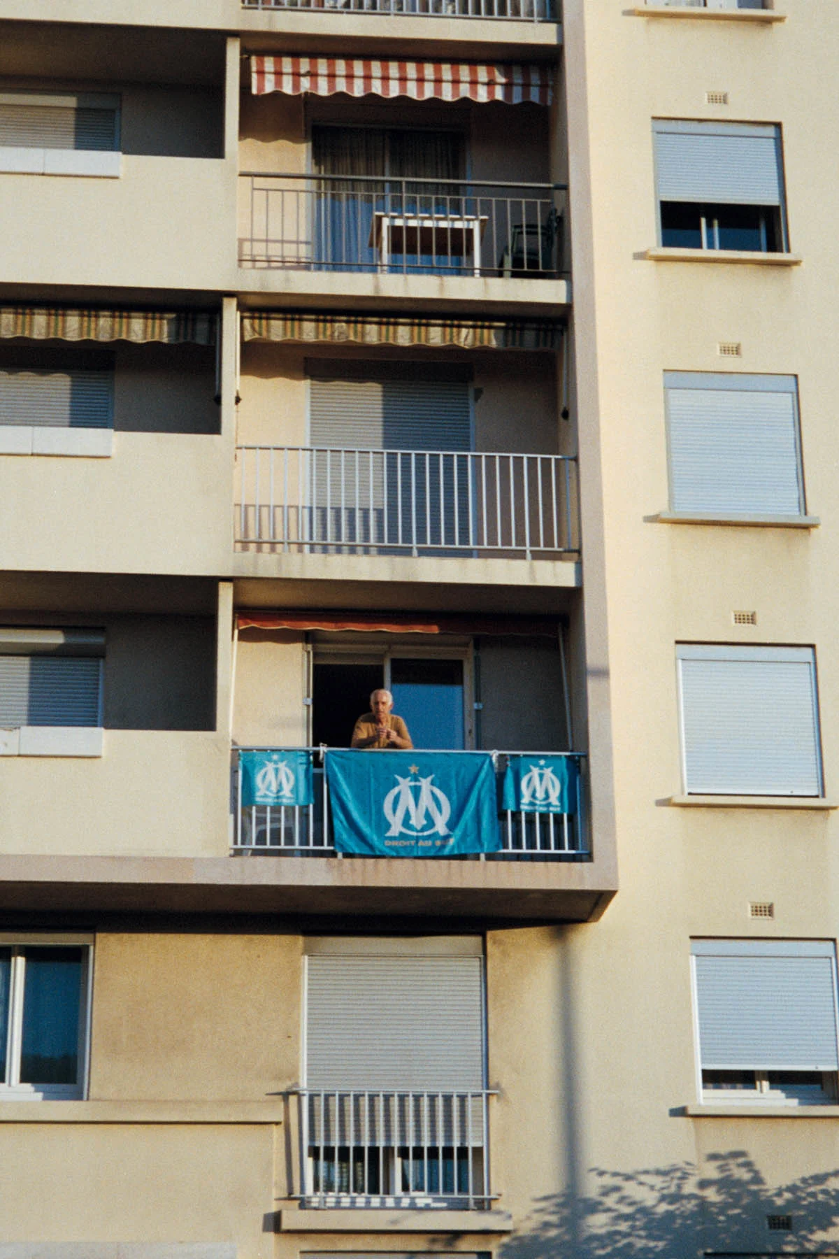 A photograph of an elderly man leaning on the terrace of his apartment hanging flags that bear the logo of Marseille football club