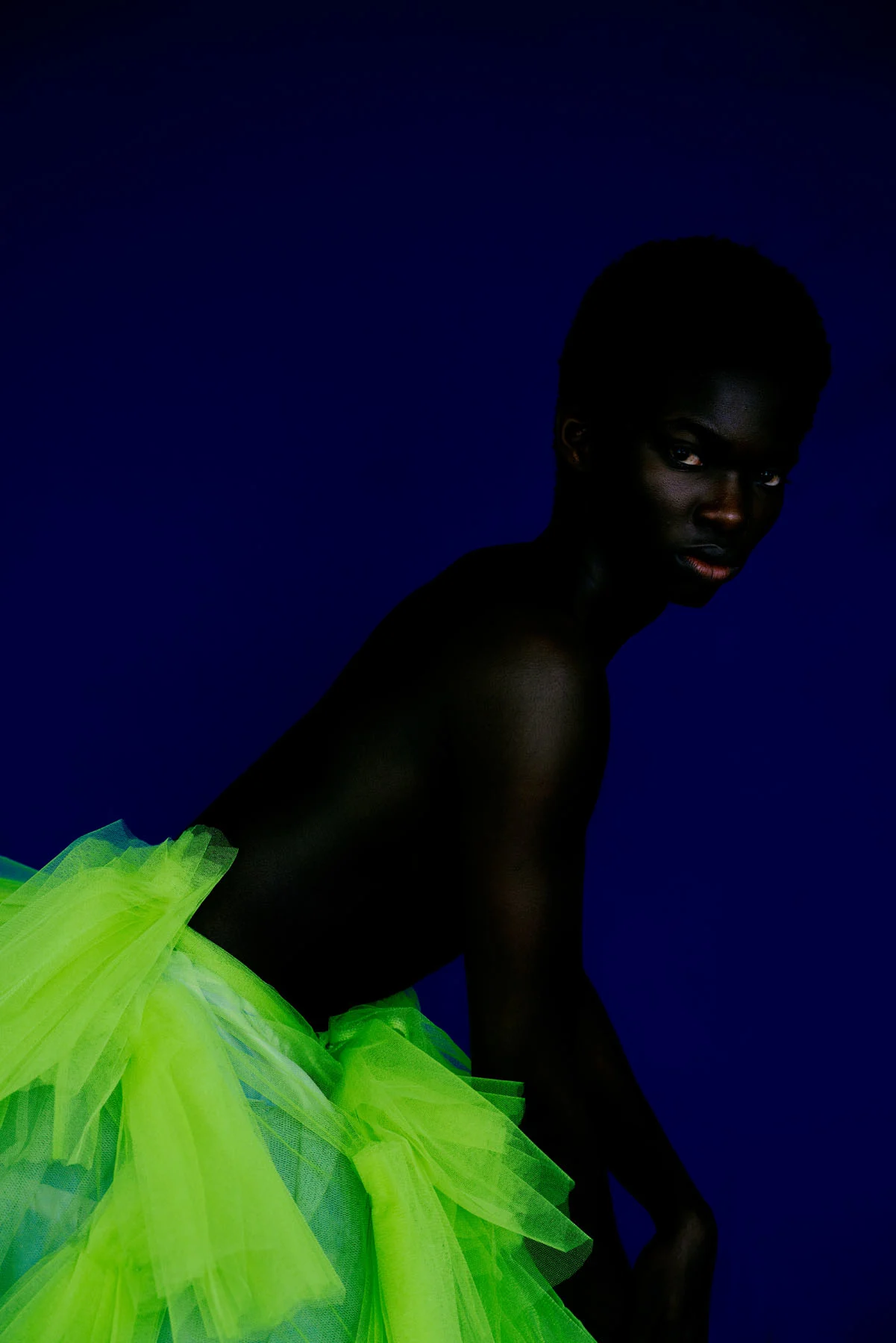 A Low-lit photographic portrait of a male model, shirtless and wearing a neon green tulle skirt, in front of a royal blue backdrop. Body profile, leaning towards the right and eyes to the camera 