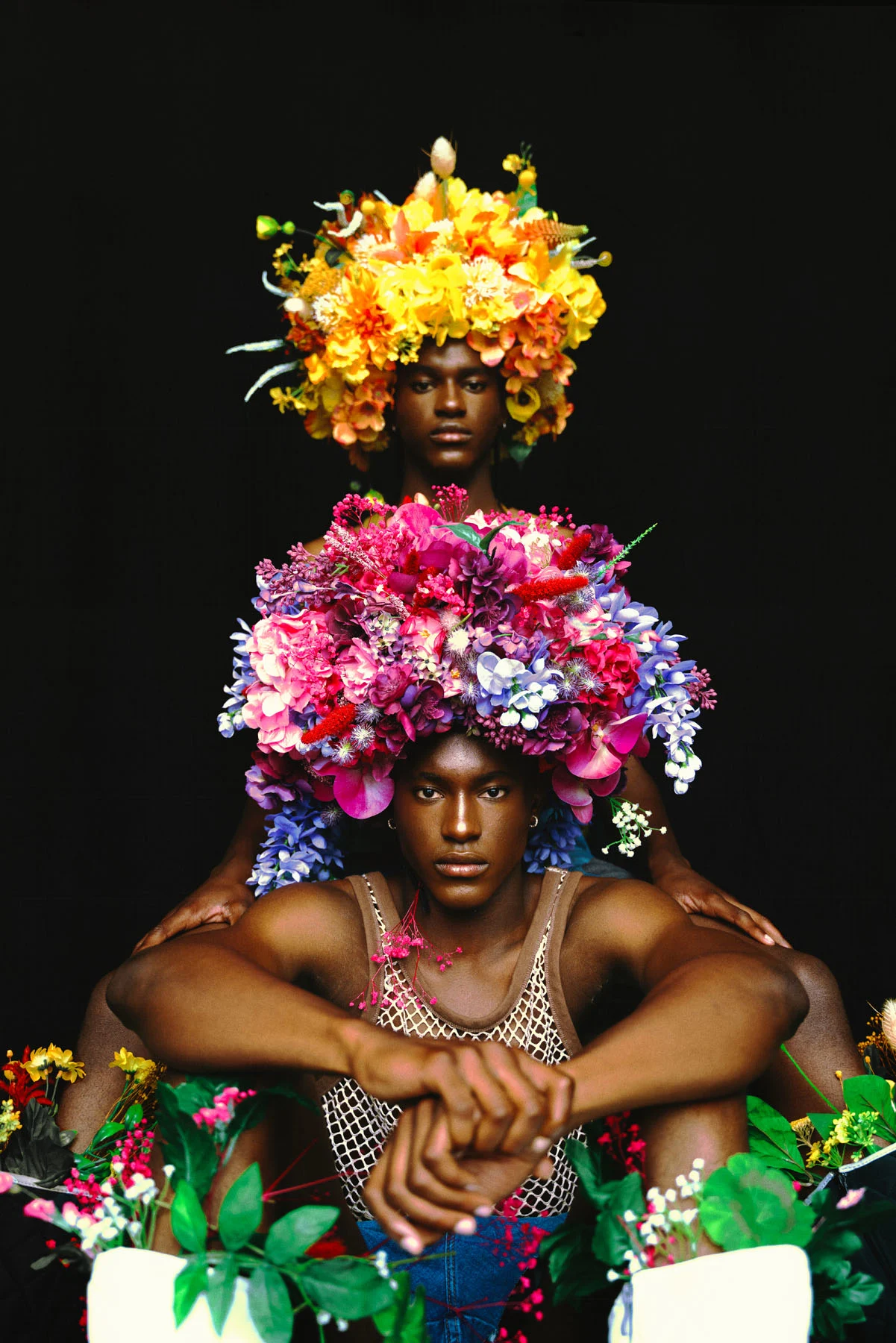 A photograph of two male twins wearing flower headpieces shaped like afros, one sitting in front of the other, and both looking into the camera.
