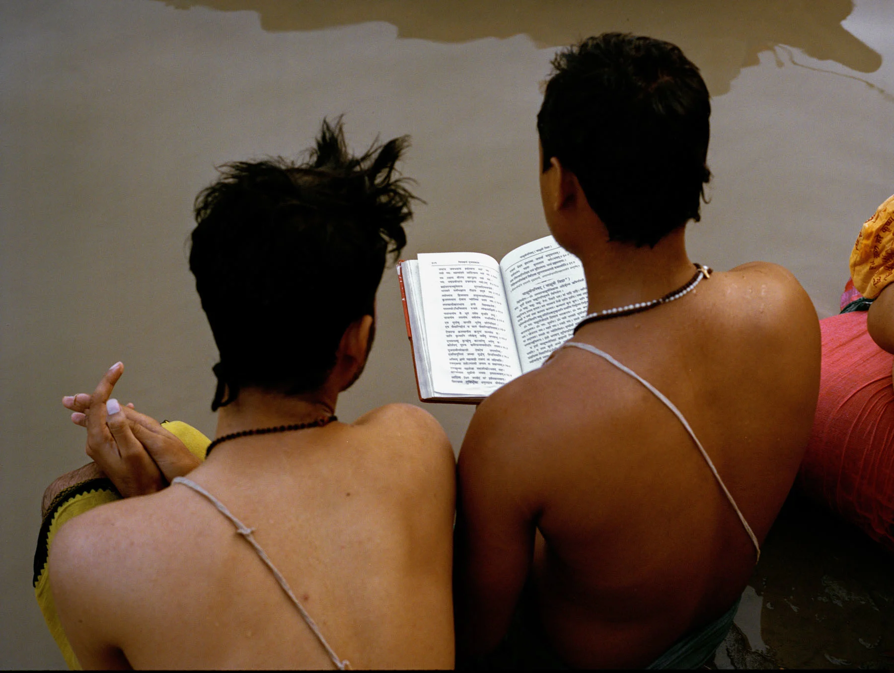A photograph of two young boys sitting by the ghat, with their legs submerged in river Ganges. They are reading the Bhagwat Gita. They are bare chested and are seen wearing a thread across their left shoulder (referred as ‘Janoi’). 