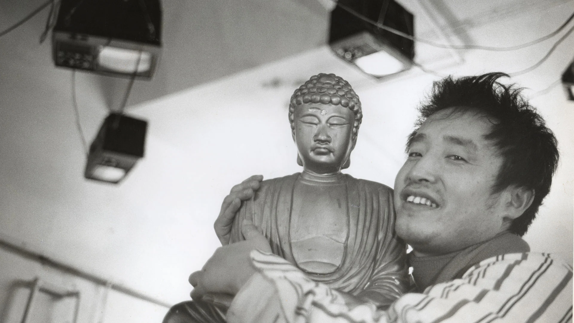 The artist Nam June Paik, an East Asian man with dark hair and wearing a white striped shirt, is pictured in a black and white photograph smiling and holding a large statue of Buddha. Three television sets or monitors are shown suspended from the ceiling behind him with cables running between them.