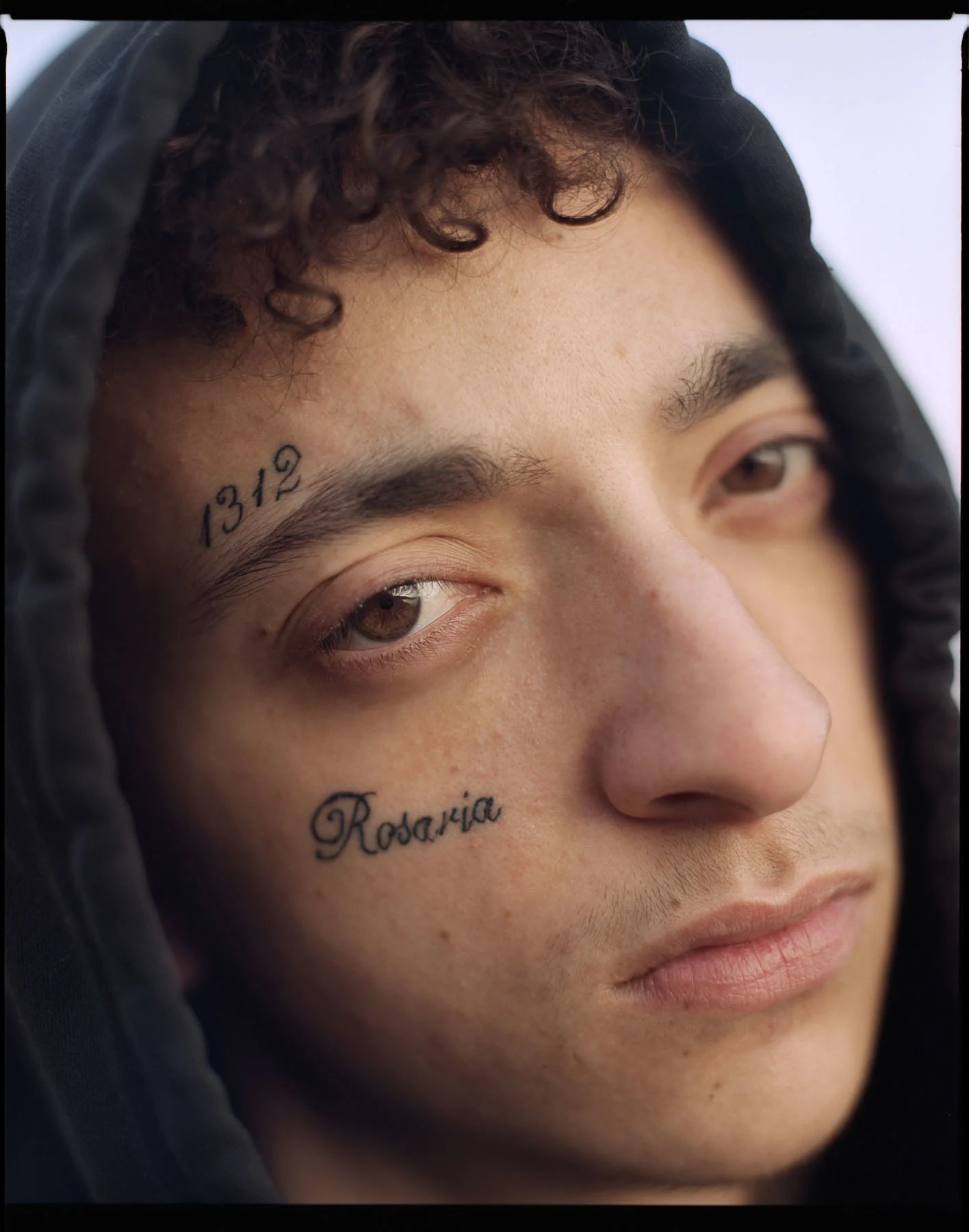 A photo of a teenage boy staring at the camera with his hood up, two tattoos around his right eye