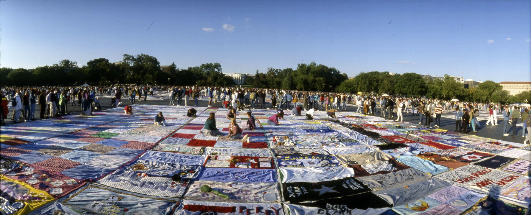 A wide color photograph showing a wide view of dozens of colorful, hand-painted quilts on the grass in Washington, D.C. Around and on the quilts stand dozens of people. Behind them, a forest of green trees is visible.