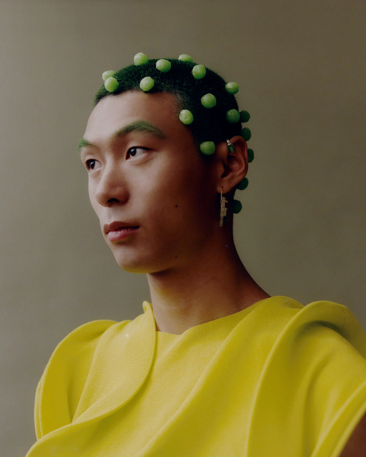 A photograph taken in the studio of model Yan Li wearing a bright yellow top with green balls attached to ther head, styled and groomed to mimic the Chinese “Guava Candy”. 