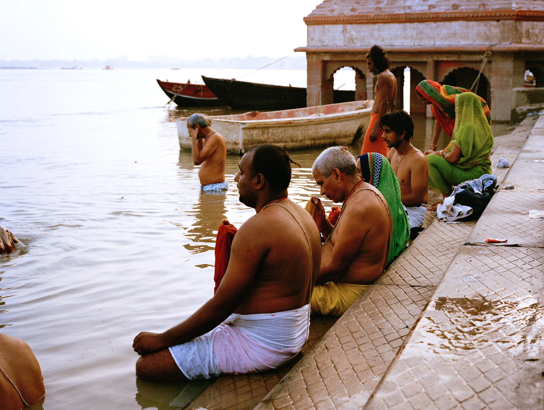 A photograph of a group of people sitting and meditating by the river Ganges, their lower halves submerged in the water. Boats are anchored by the river in the background. 
