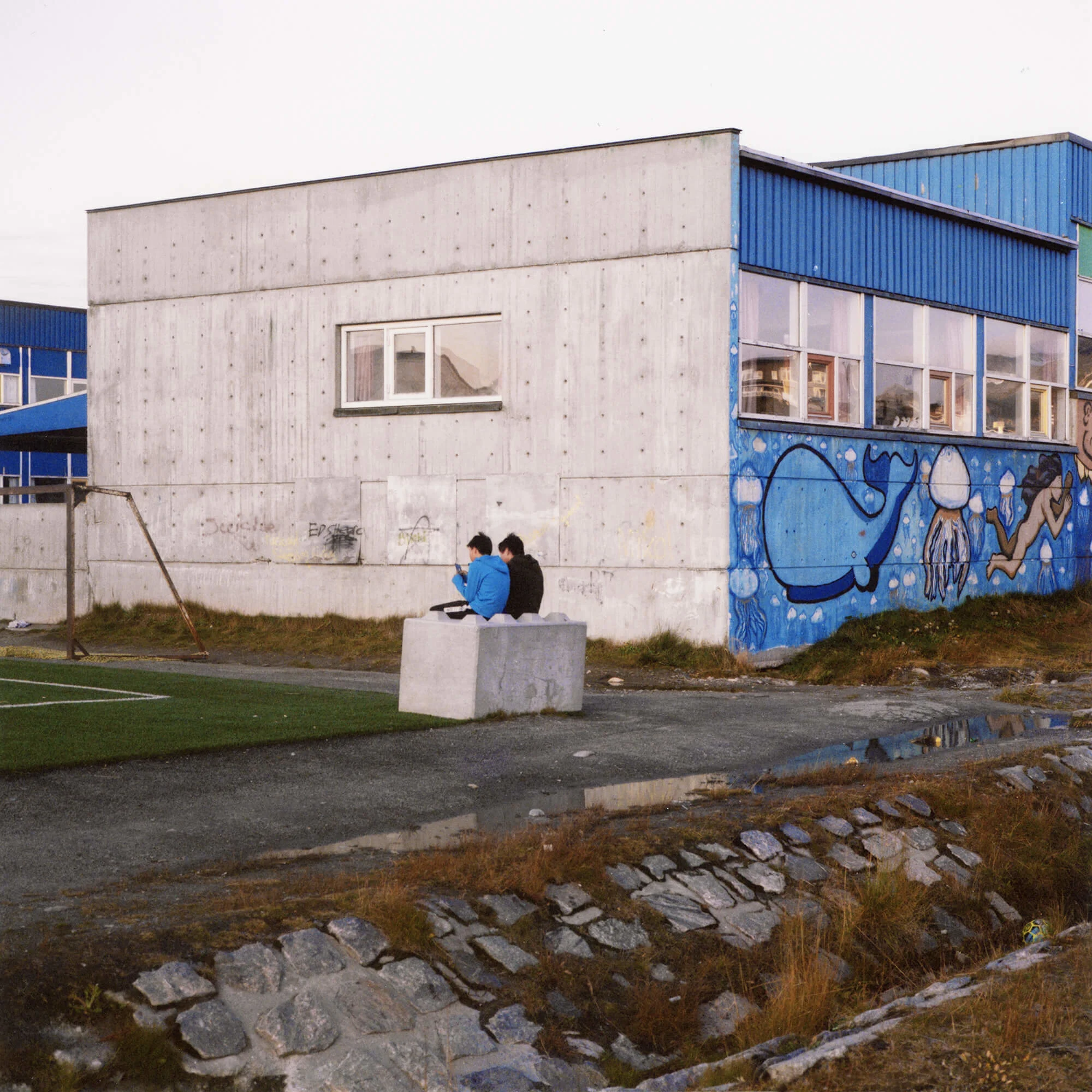 Two friends on their phones hanging out on the football court in Nuuk. | Boys playing after school, in Nuuk.
