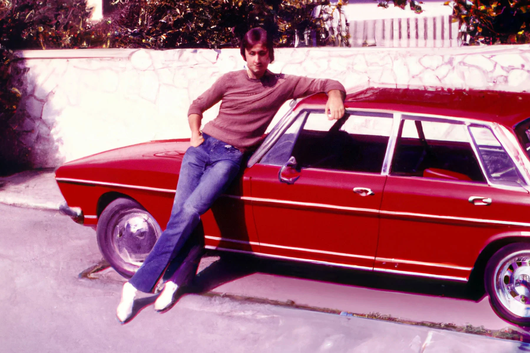 An image that looks like a photograph but is created with the use of Artificial Intelligence. The image has an 80s aesthetic, showing a man around 25-30 years old leaning proudly on a shiny red car parked outside what looks like a stone fence of a house. 