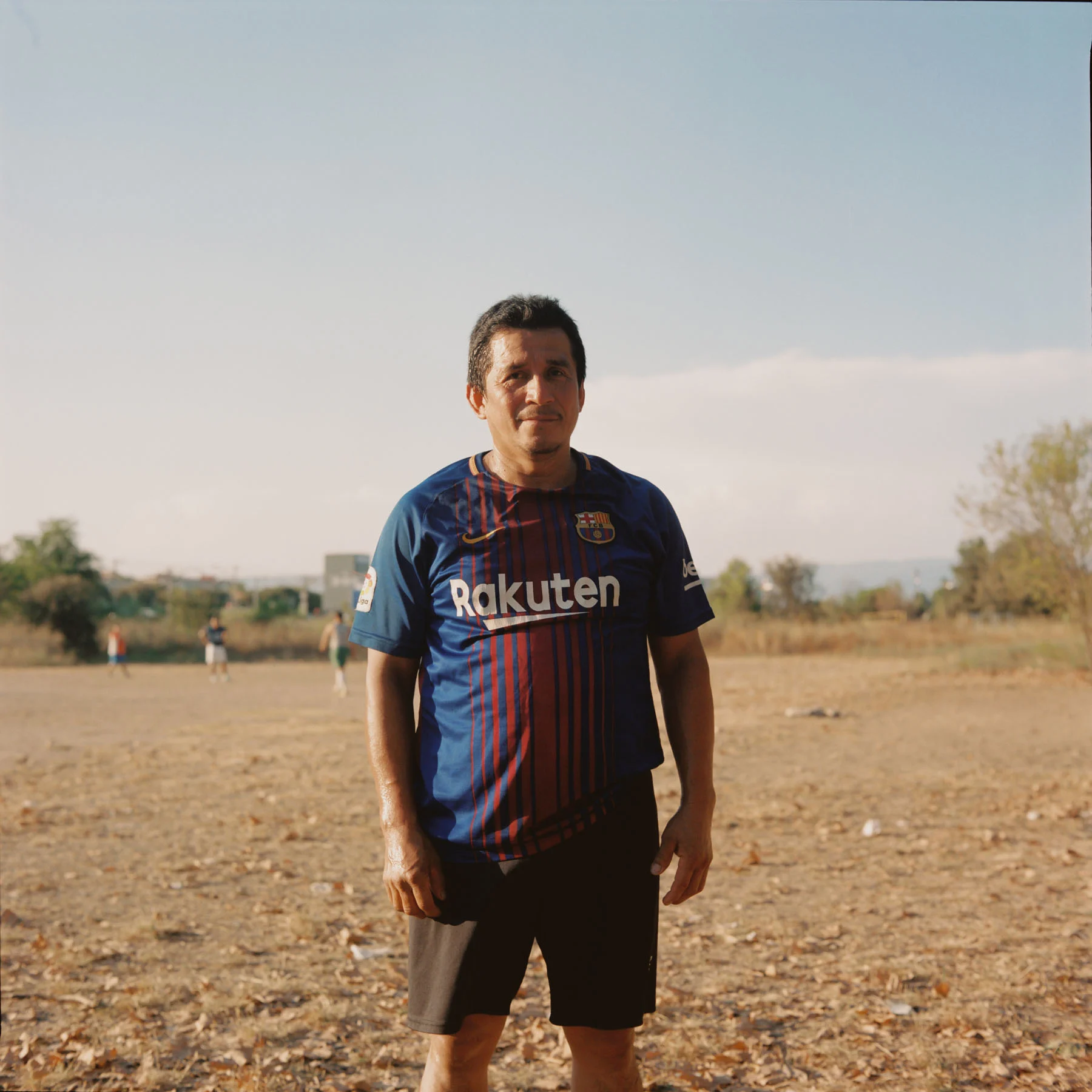 A photograph of a man posing on a soccer field. The man is wearing an FC Barcelona soccer jersey. There are other players out of focus playing in the background.
