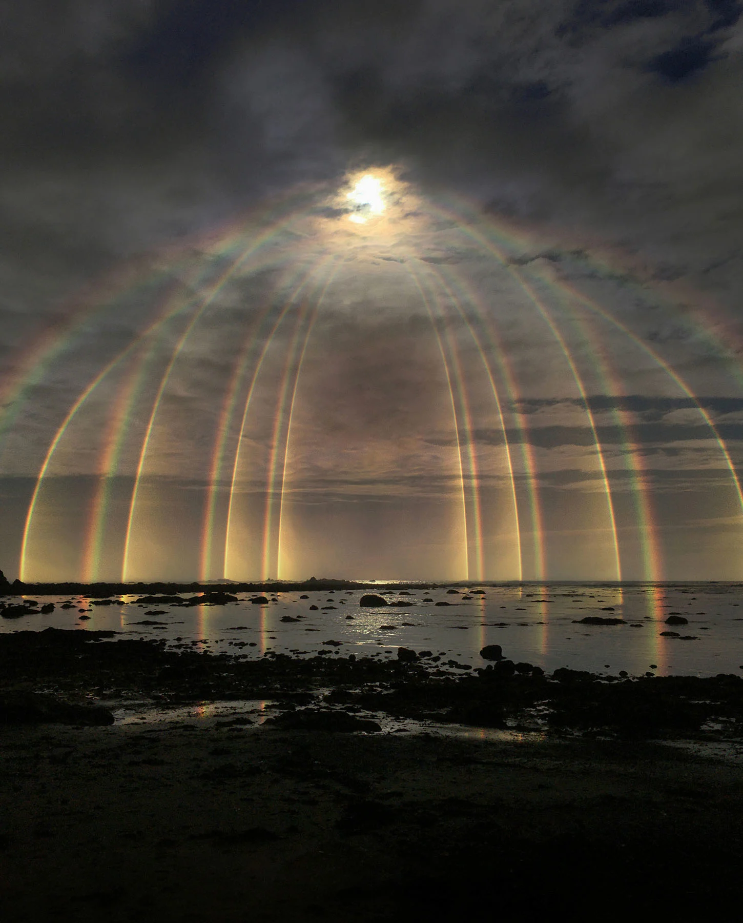 A photograph of a partly flooded landscape covered prominently by a cascading gate of rainbows. The rainbows radiate downward from the sun in the sky, towards the flooded horizon line. 