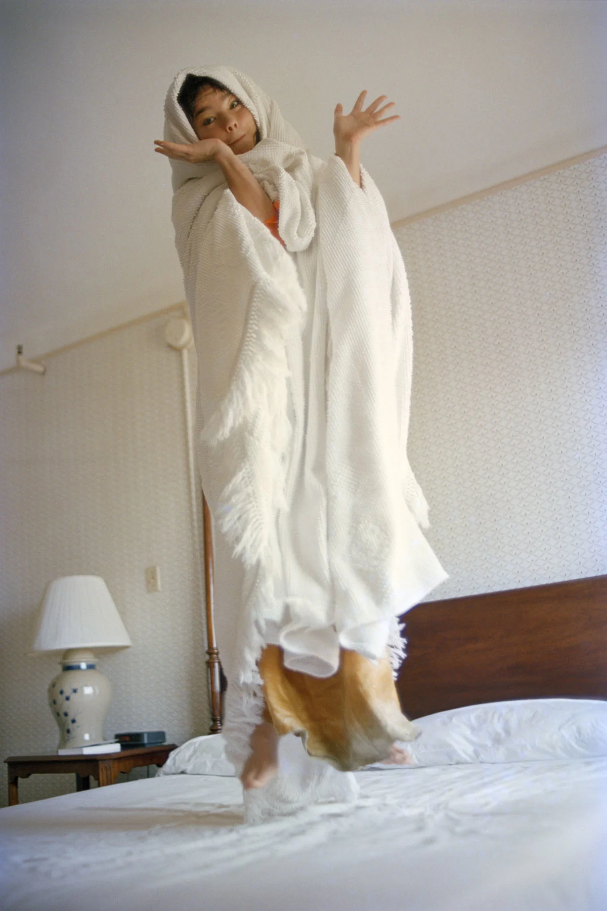 Photograph of the musician Björk, a young, dark-haired woman wrapped in a white bed sheet, jumping on a hotel bed with her hands spread out to the sides. 