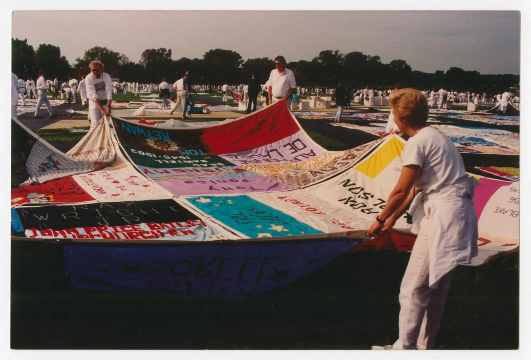 A color photograph of colorful hand-sewn quilts being laid down on the grass by three figures in the foreground. All wear white. 