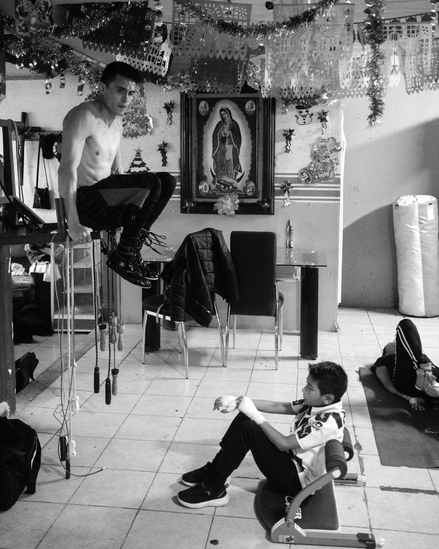A black-and-white photograph of two boxers training with religious sculptures and the image of the Virgen de Guadalupe in the background. 
