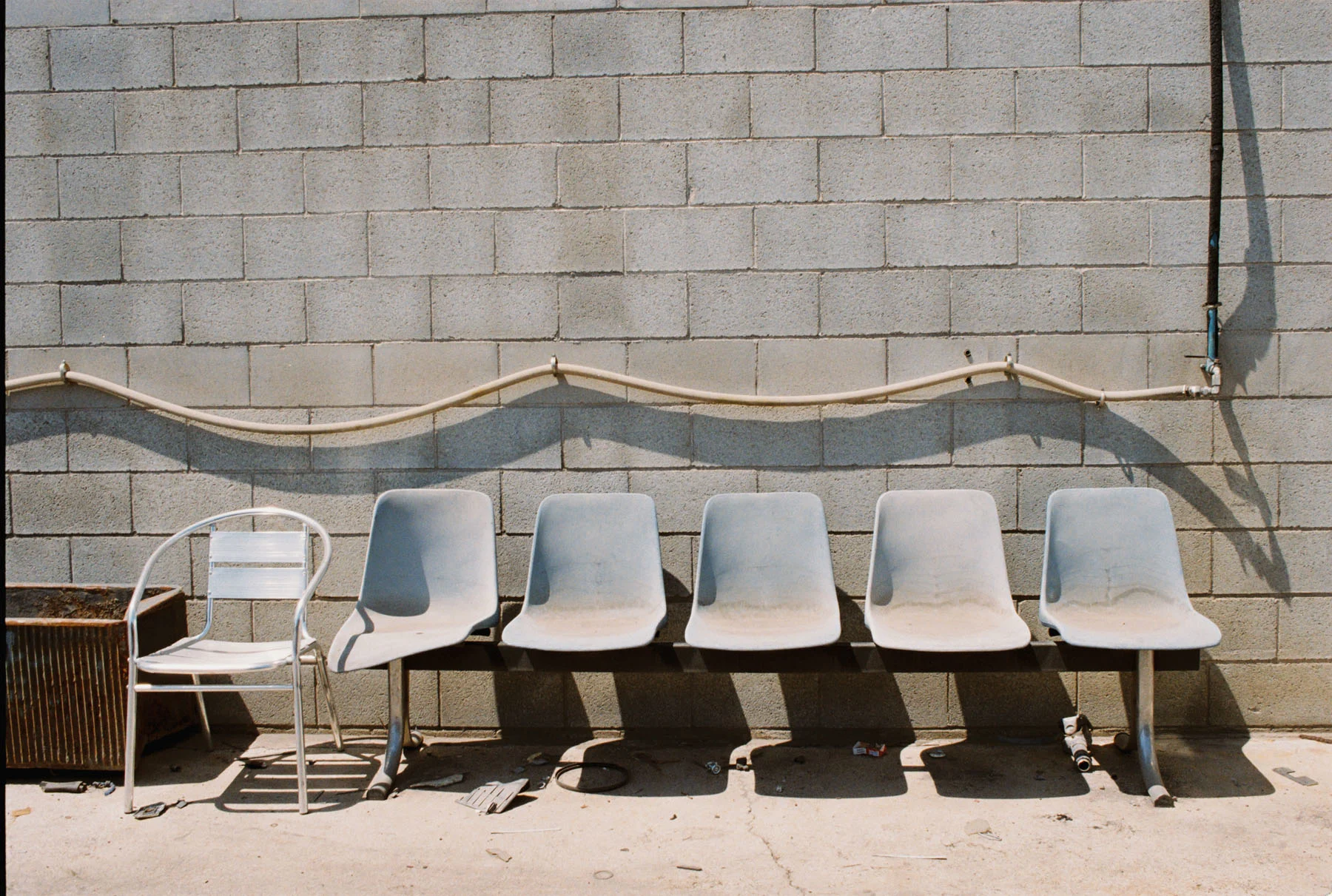 A photograph of five blue chairs laying in an empty backyard. Another metal chair sits beside them. In the background is a grey brick wall with pipes and hoses attached to it.
