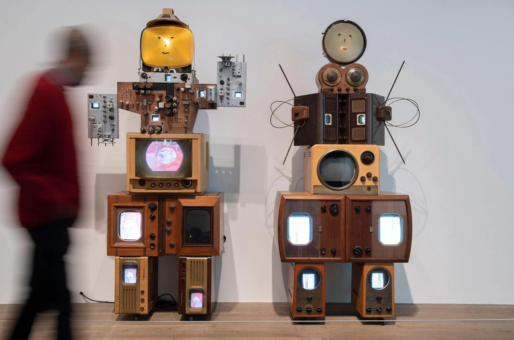 Two sculptures, shaped like robots, stand next to each other in front of a white wall at a museum. Each sculpture is made up of a number of old televisions and switchboards. Both appear to have faces. A blurry museum goer on the move is visible to the left.