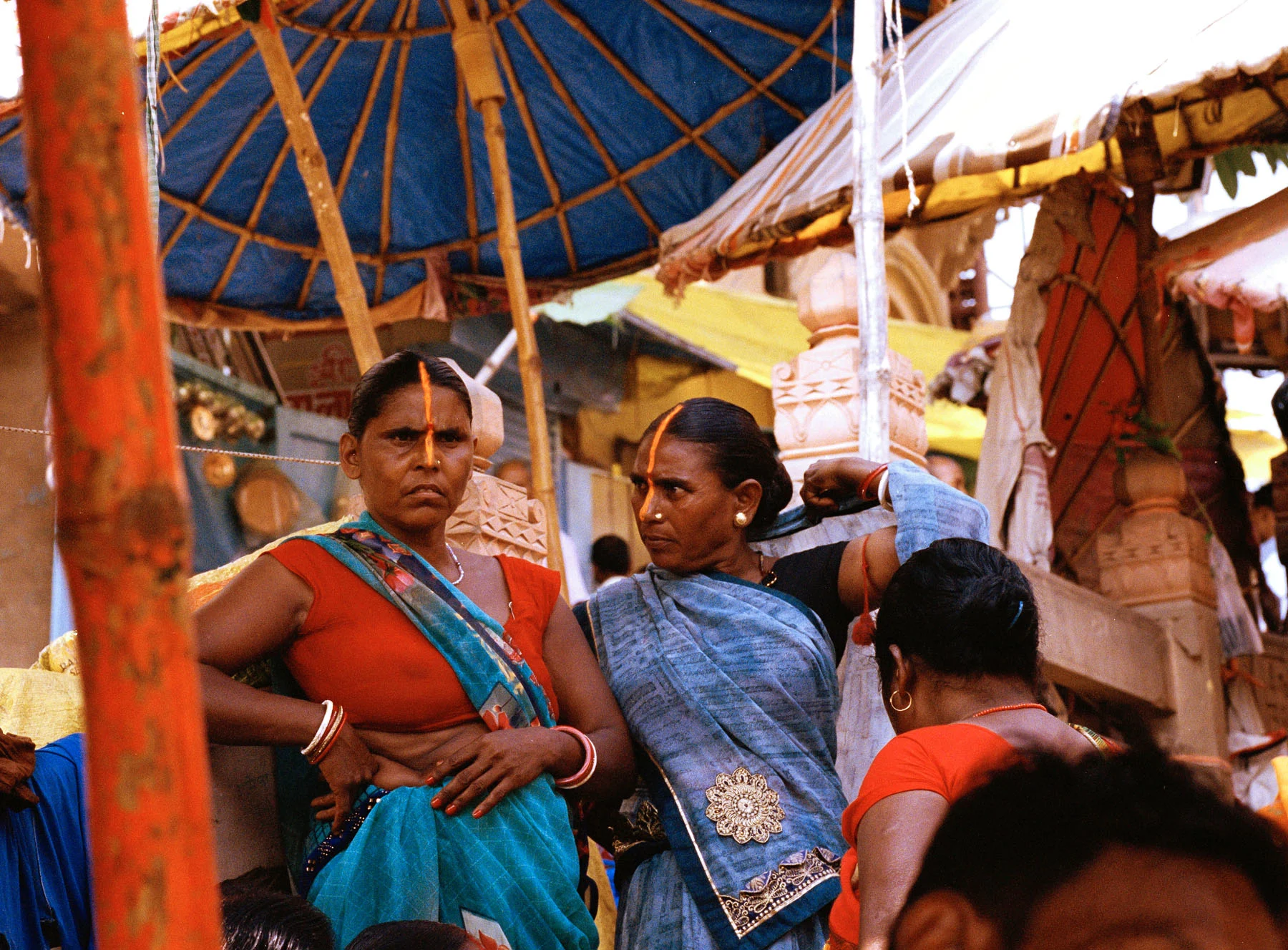 A photograph of two women wearing sarees and their faces adorned with Sindoor (red vermillion powder stretching from the tip pf the nose to the center of the head) signifying their marriage.