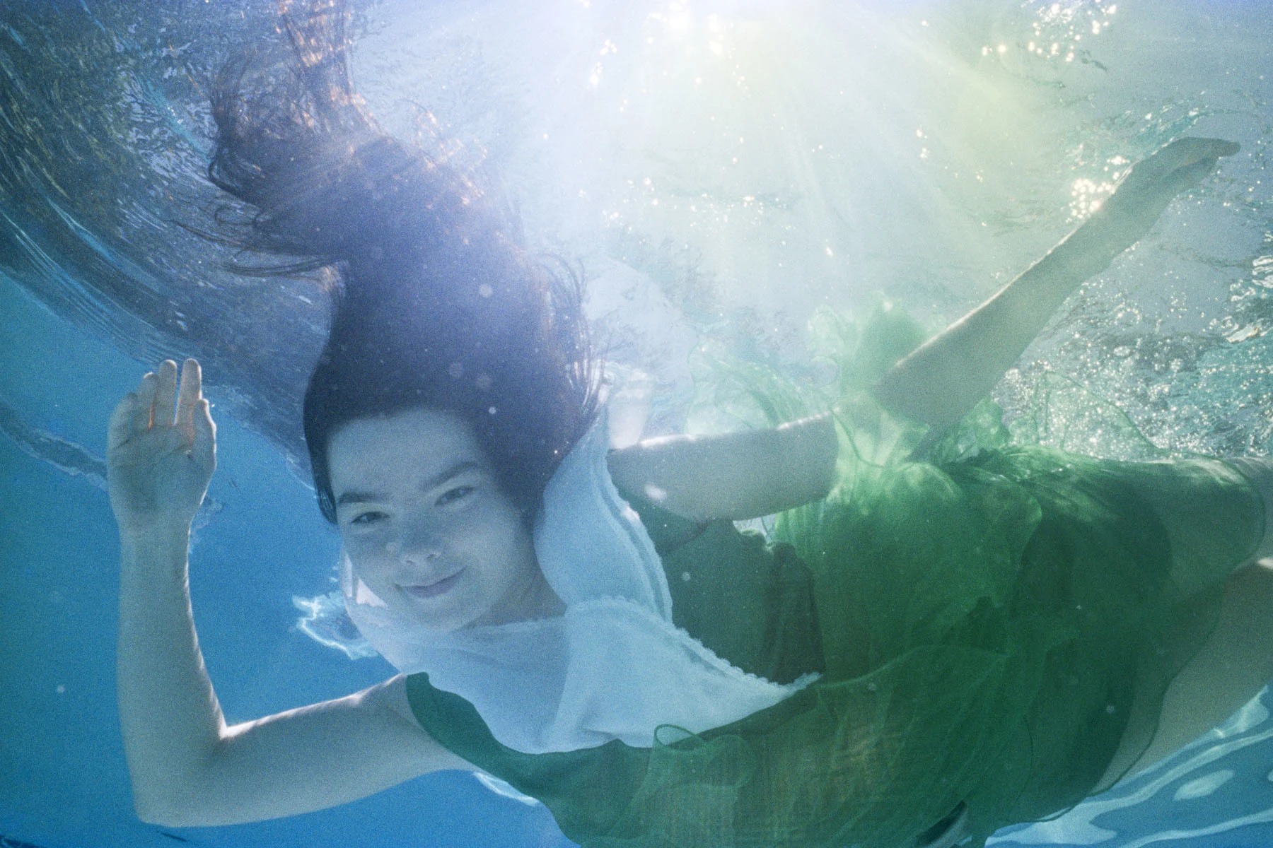 Photograph of the musician Björk, a young, dark-haired woman wearing a green dress. She is shown beneath the deep blue surface of a swimming pool, pictured from below, waving at the camera and smiling. The sun’s rays are lighting the scene from above.