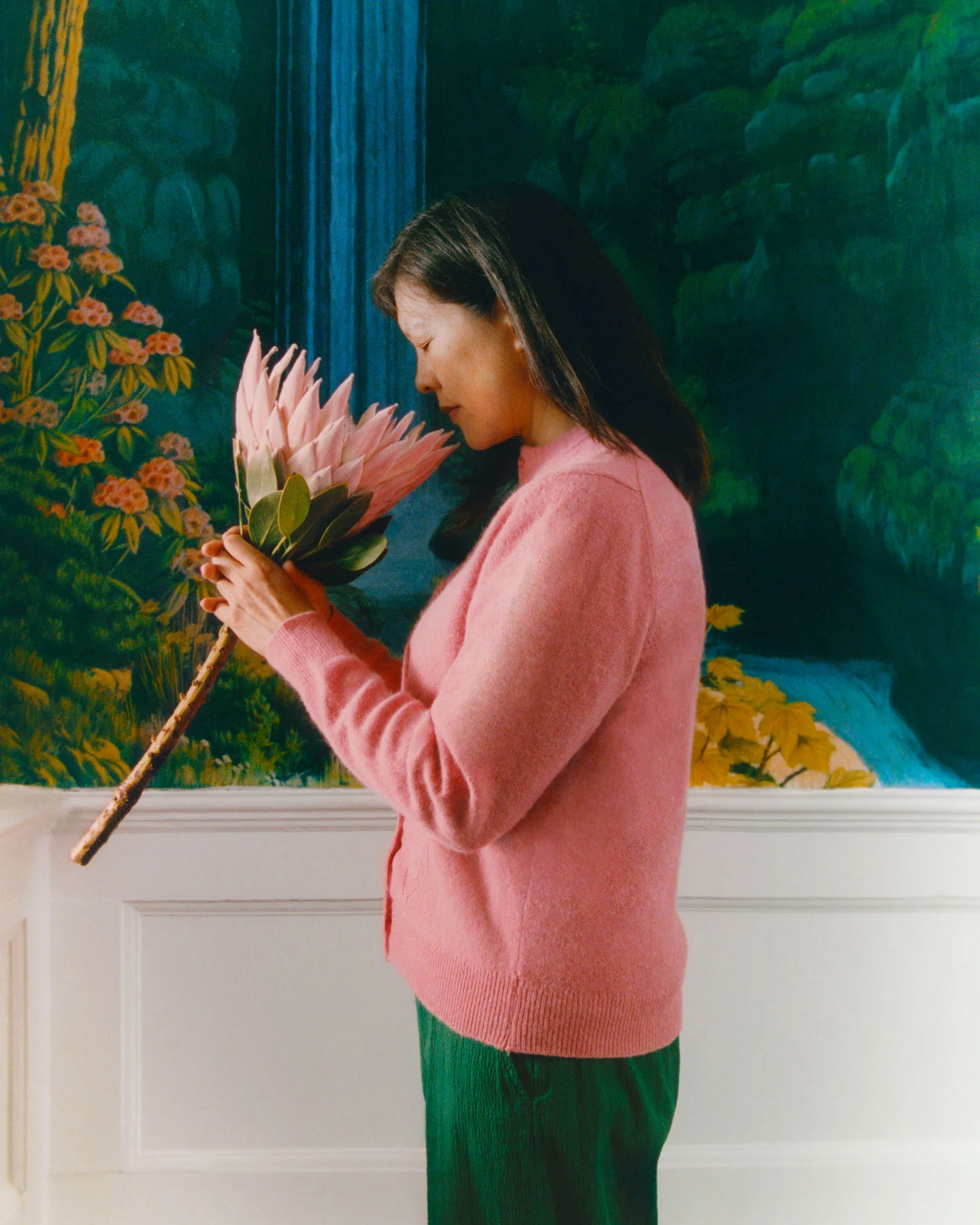 A photograph of ceramicist Sassirika Lam, shot in front of a waterfall mural of her Buddhist temple, smelling a pink protea flower.