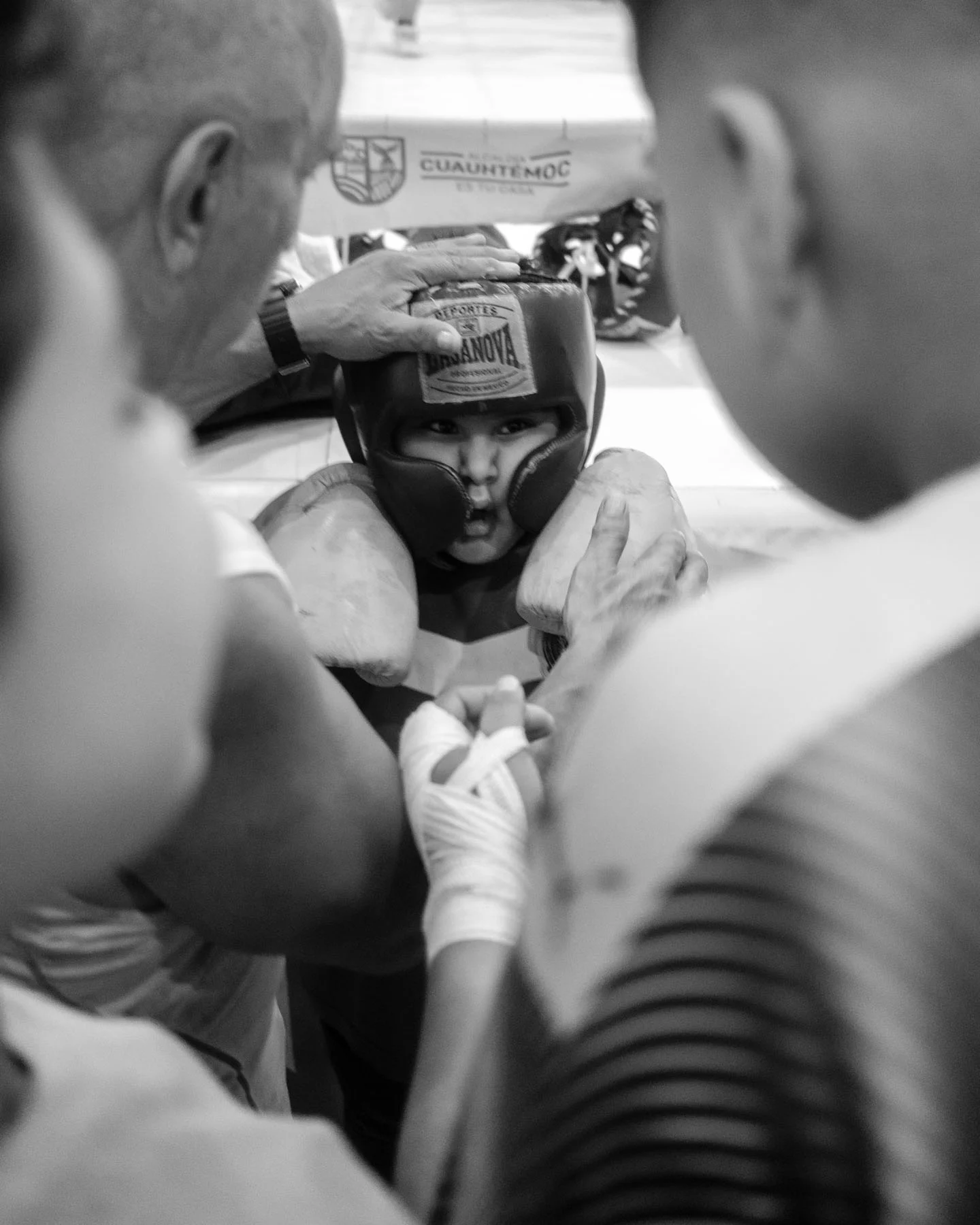 A black-and-white photograph of a young boxer preparing to enter the ring, as men adjust his helmet and offer him support.