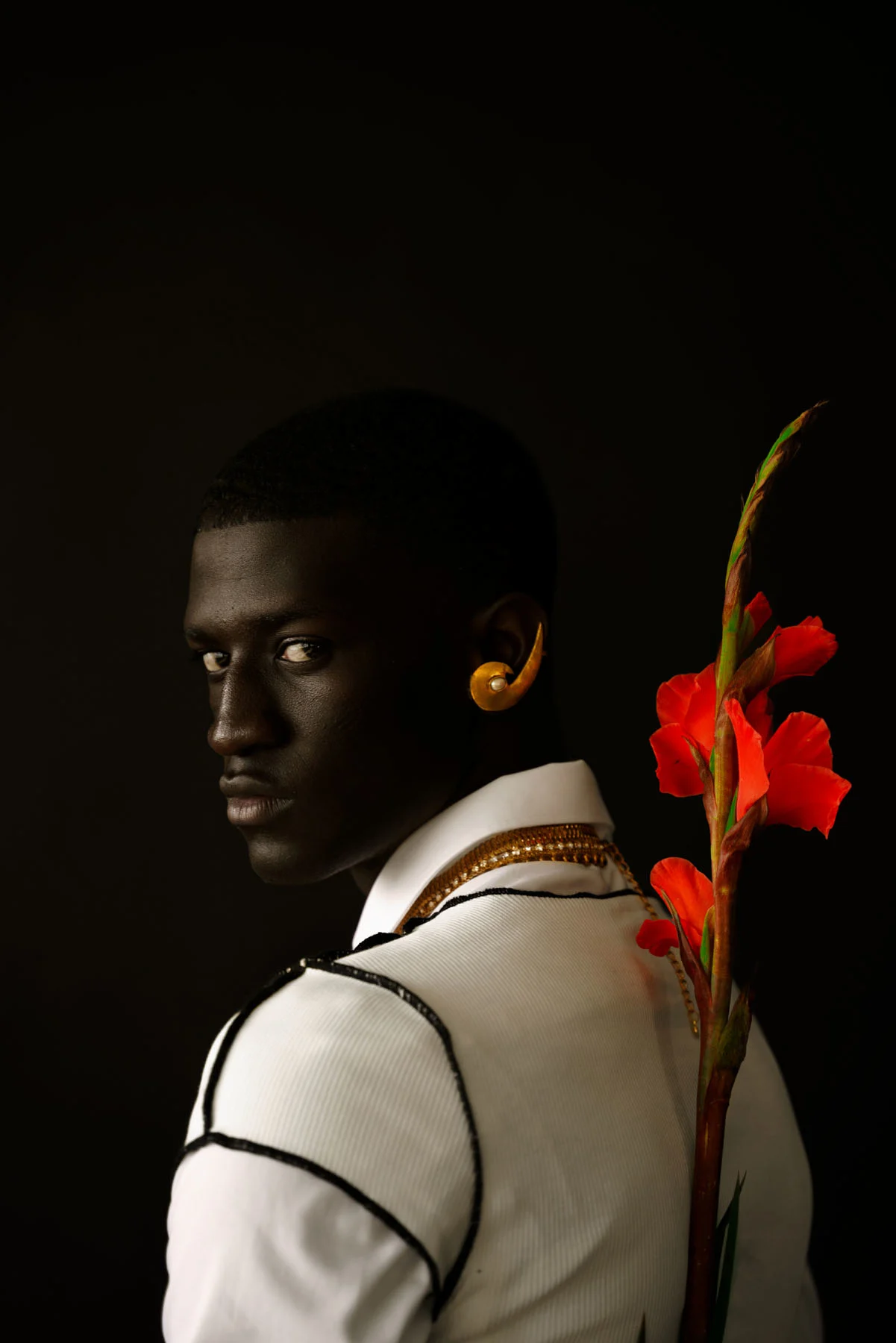 A low-lit photographic portrait of a male model wearing a collared white shirt and gold accessories, holding an orange flower in front of a black backdrop. He faces towards the camera as his body faces away from the camera.