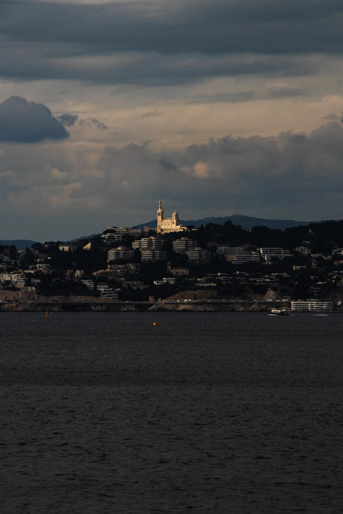 A photograph of Marseille’s Roucas Blanc hill, taken on a cloudy day facing the sea with sun lighting up the hill’s basilica