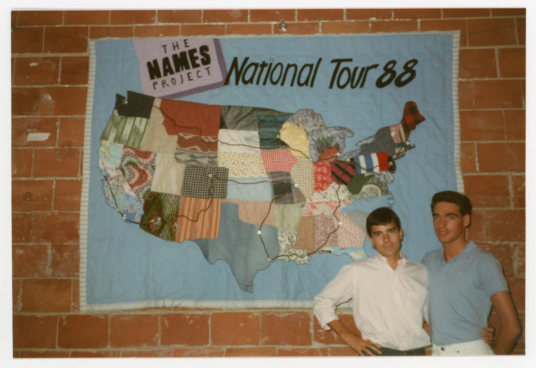 A color photograph of two men standing in front of a quilt panel that features a map of the United States with a map of stops on the Names project national tour in 1988. The text on the quilt reads, "The Names Project National Tour 88".