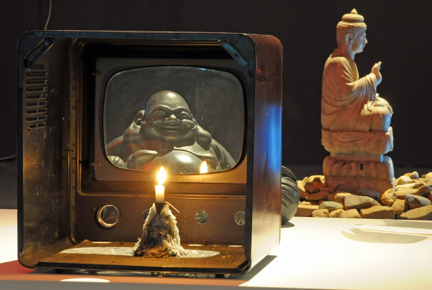 A hollowed-out television holds a burning wax candle. Visible through the screen is a small statue of a smiling Buddha. In the background, on a small pile of pebbles, is another figurine.