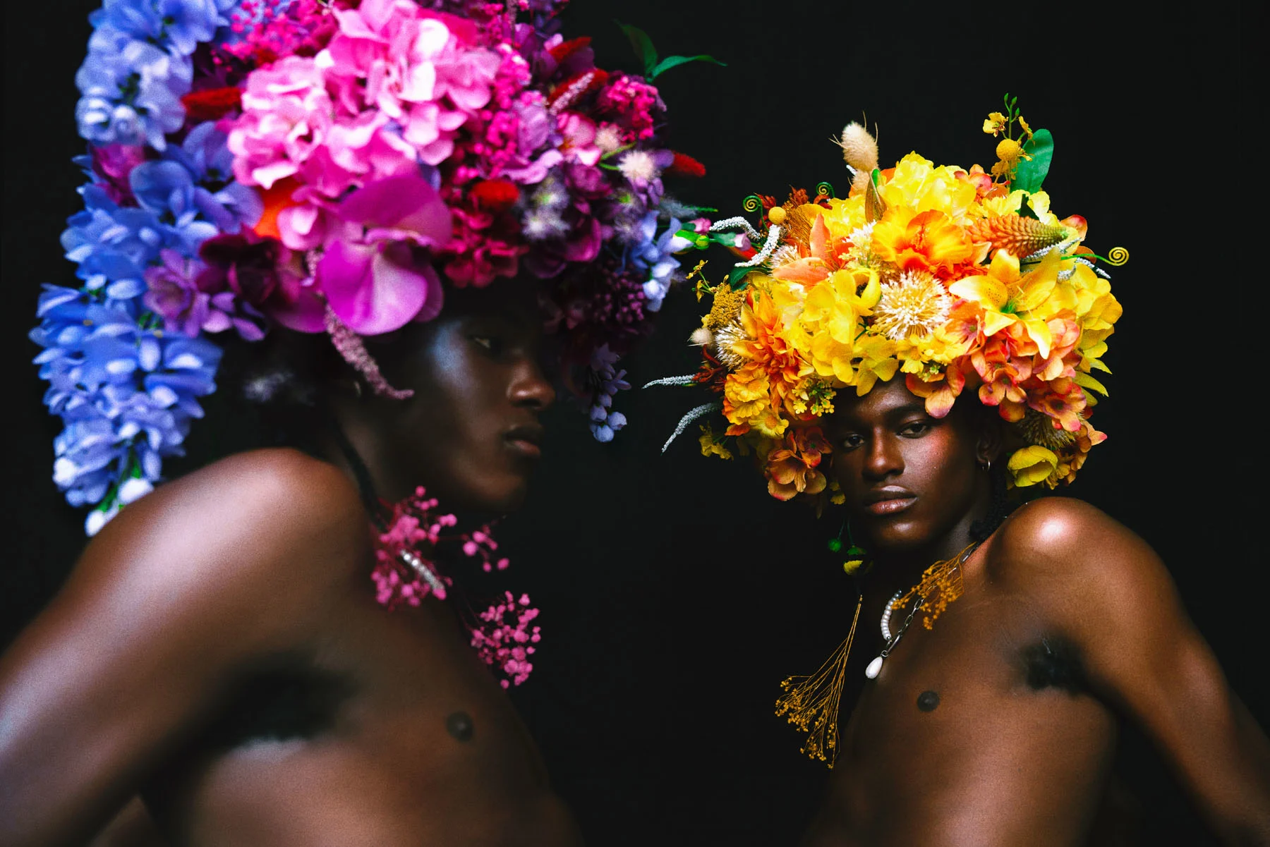 A photograph of male twins wearing flower headpieces shaped like afros, sitting across from each other. The twin with a purple headpiece looks to the right while the twin with the yellow headpiece looks at the camera.