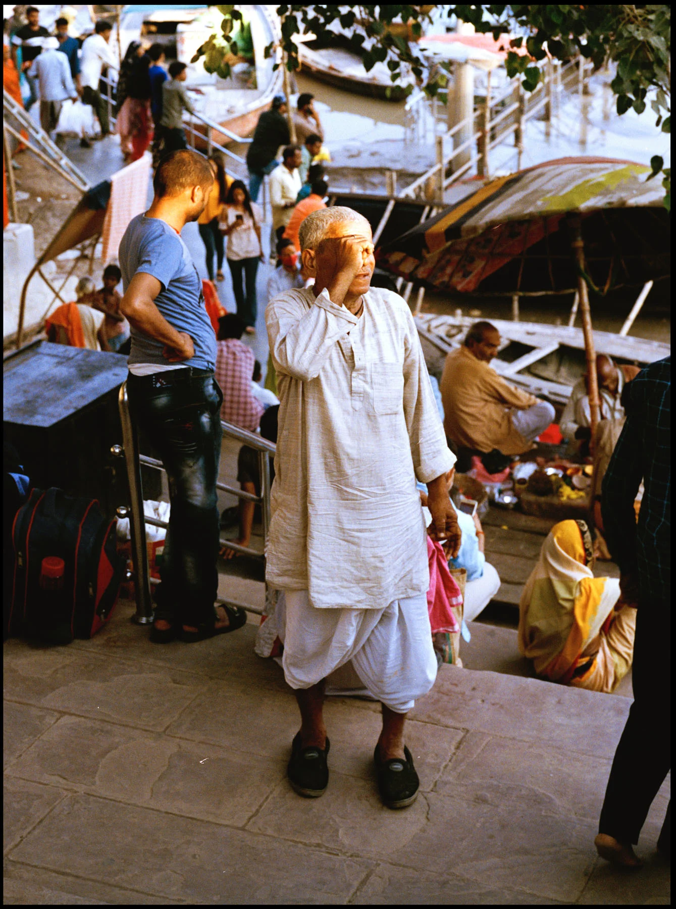 A photograph of an old man wearing white Kurta and Dhoti pants, captured on the steps of the ghat (the series of steps leading down to a body of water or wharf). There are other people at the ghat, in the background of the photograph.