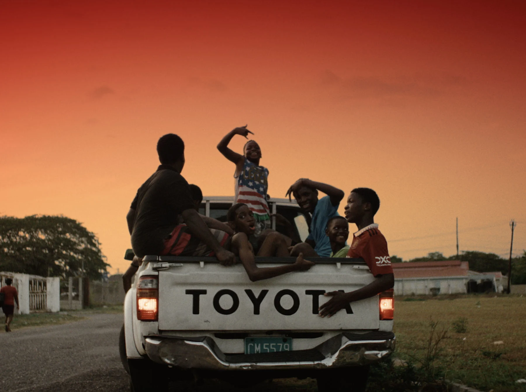 A still from the film "Blue Eyes" by Andre Muir, showing a group of kids sat in the back of a Toyota truck, posing towards the camera. 