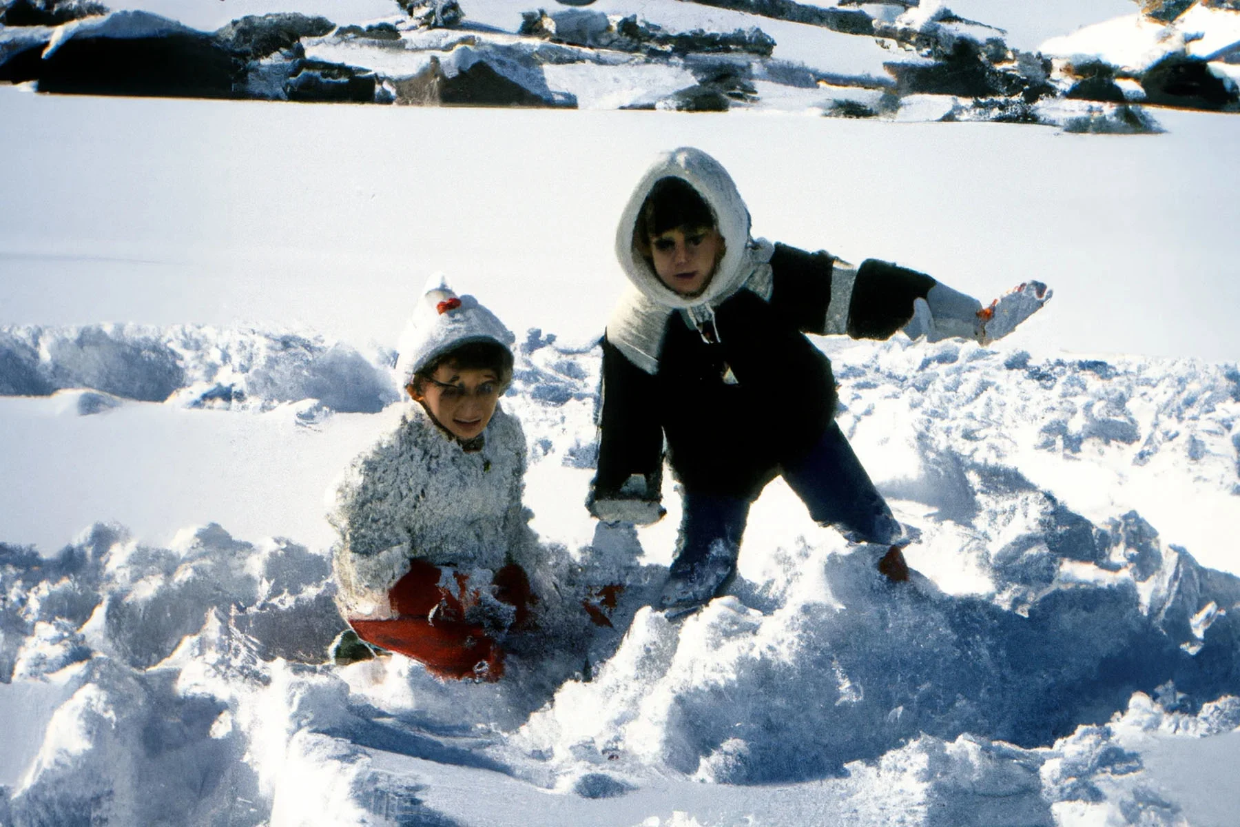 An image that looks like a photograph but is created with the use of Artificial Intelligence. The image has a 90s feel, showing a candid scene of two young kids playing in the snow on a sunny day, dressed in warm clothes and hats, looking at the camera.