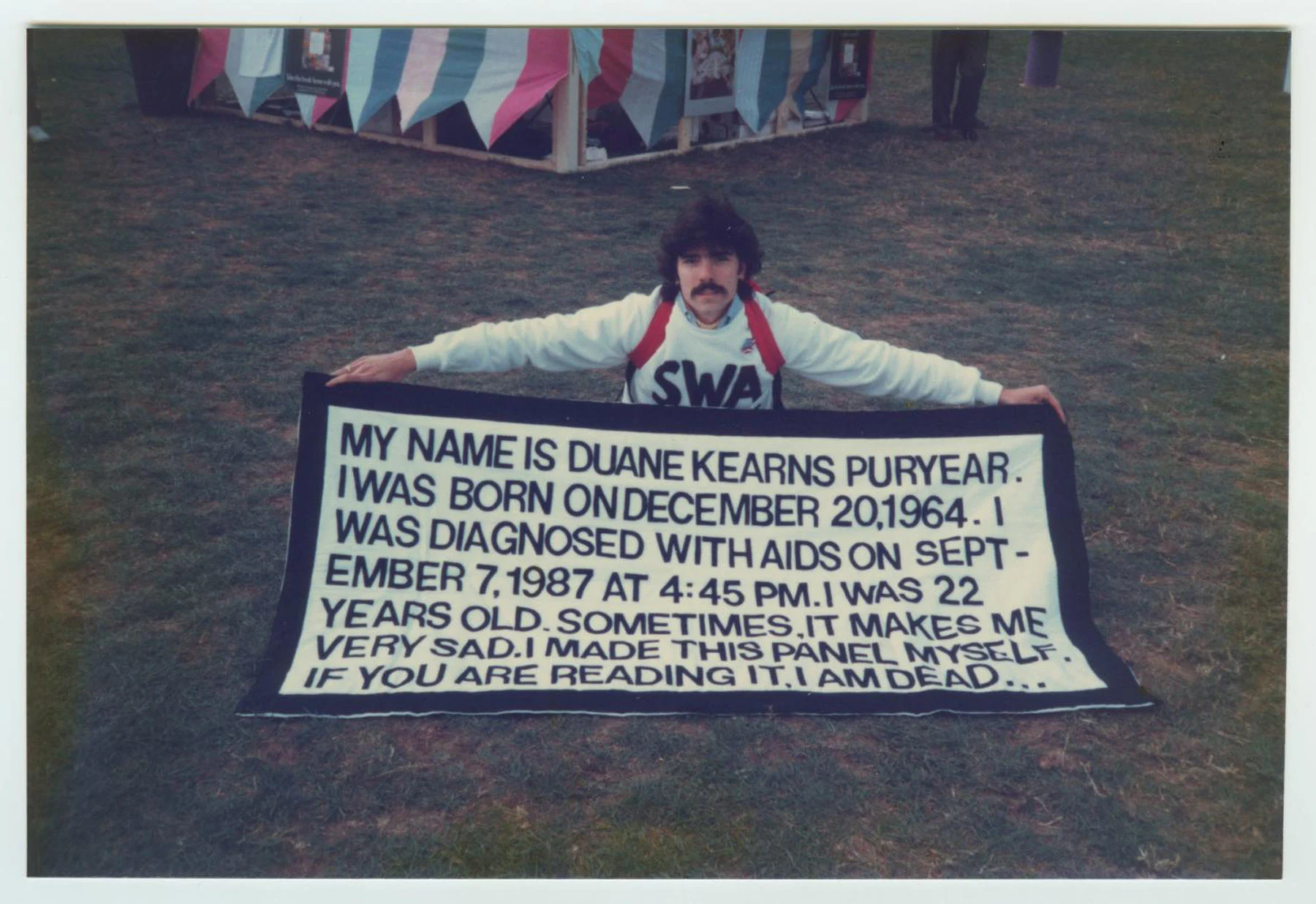 A color photograph of a man wearing a white crew neck with the letters “SWA” across it in black kneels on the grass. He holds a wide black-and-white sign that reads, in black letters on a white background: “MY NAME IS DUANE KEARNS PURYEAR. I WAS BORN ON DECEMBER 20, 1964. I WAS DIAGNOSED WITH AIDS ON SEPTEMBER 7, 1987, AT 4:45PM. I WAS 22 YEARS OLD. SOMETIMES IT MAKES ME VERY SAD. I MADE THIS PANEL MYSELF. IF YOU ARE READING IT, I AM DEAD…”