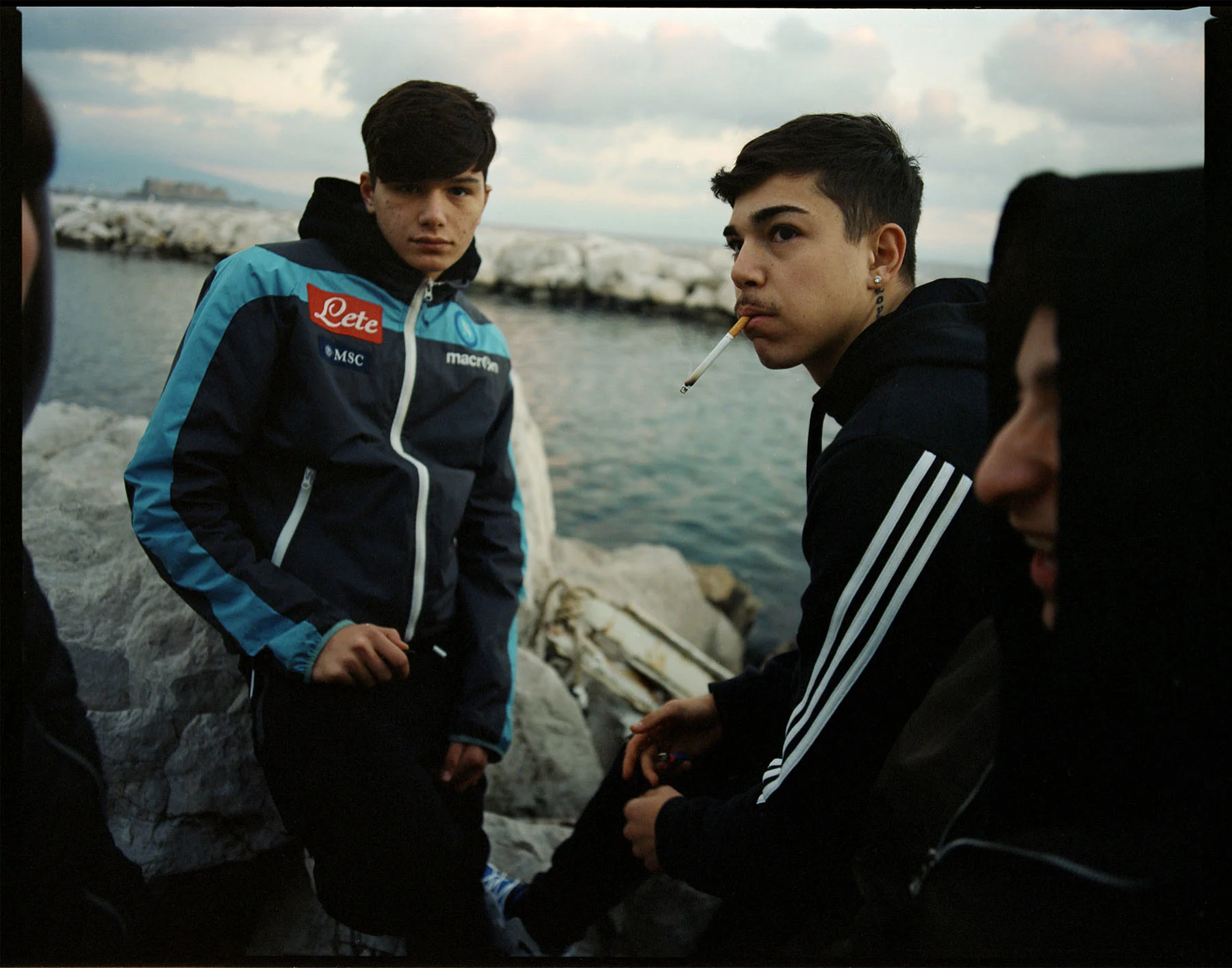 A photo of a group of teenage Neapolitan boys hanging out by the sea, a cigarette hanging from one of their mouths