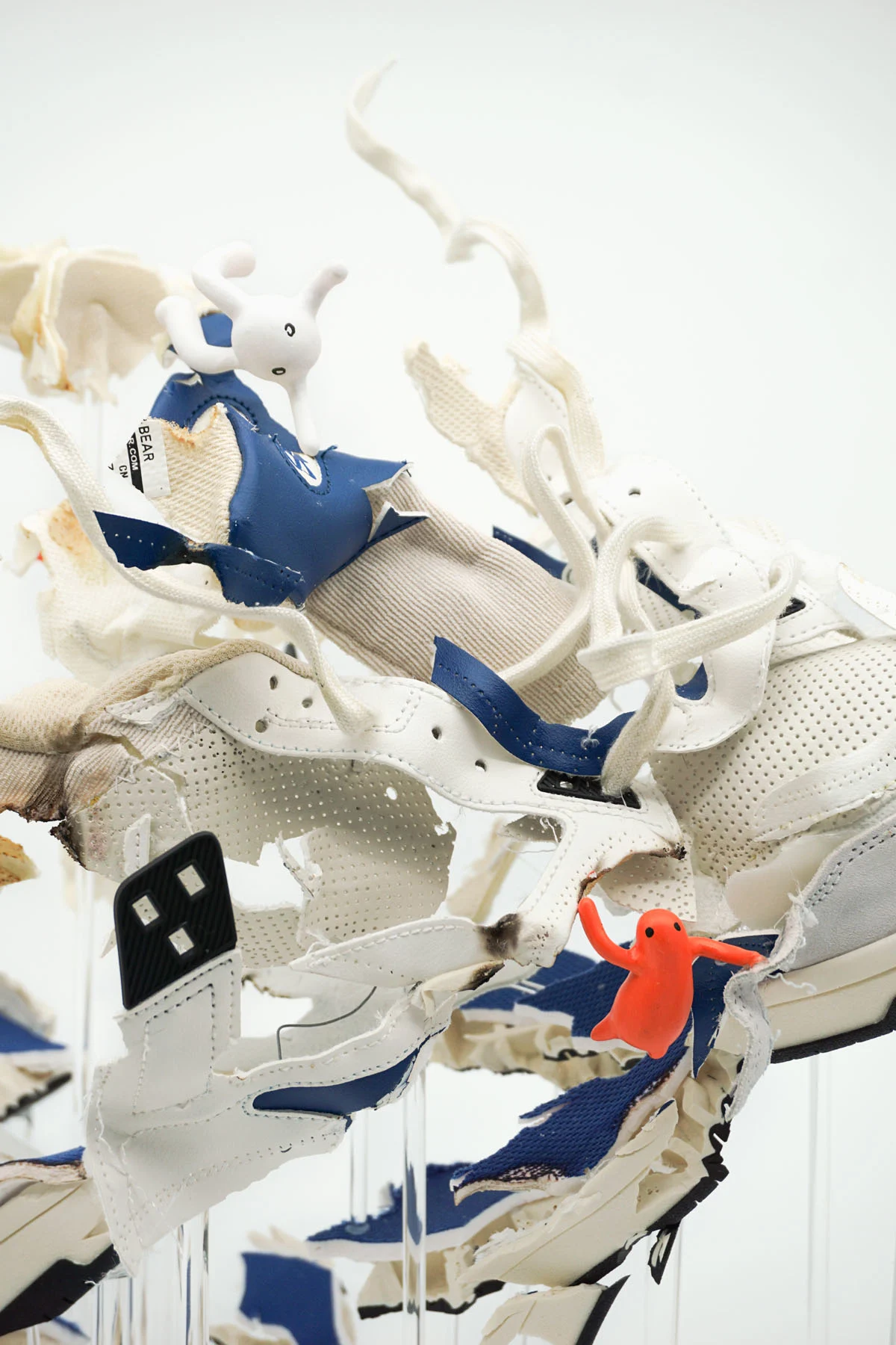 A close-up photograph of a sculpture made from pieces of sneakers. The sculpture shows a sneaker that looks like it's exploding, with small orange figurines hanging on to various pieces.  