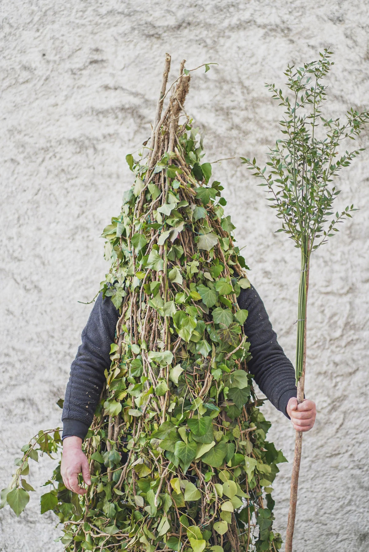 A photograph of a man wearing a “Rumita” outfit (made from tree branches) and holding a stick called a “fruscio” while standing in front of a white wall in Satriano village, Italy.