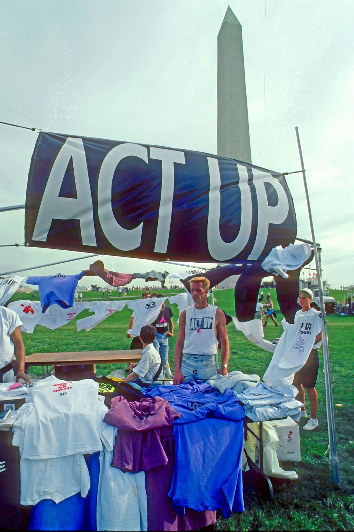 A color photograph of a man wearing a sleeveless white shirt with the black ACT UP logo across his chest. He stands behind a table laden with T-shirts, under a large “ACT UP” sign. The Washington Monument is visible behind them.